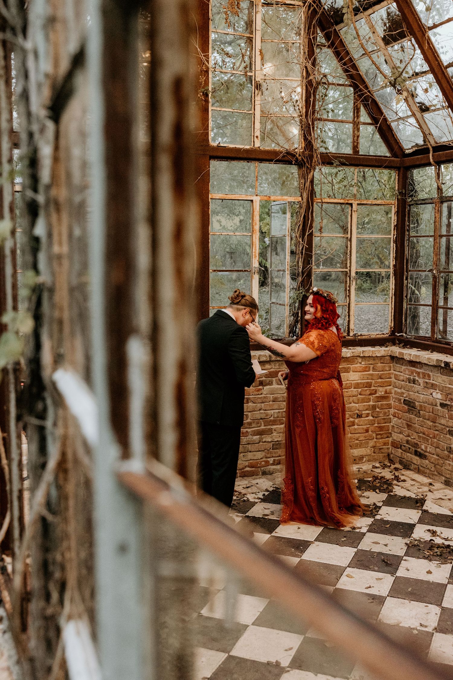 A couple shares an intimate moment inside a vine-covered abandoned greenhouse with checkered floors.