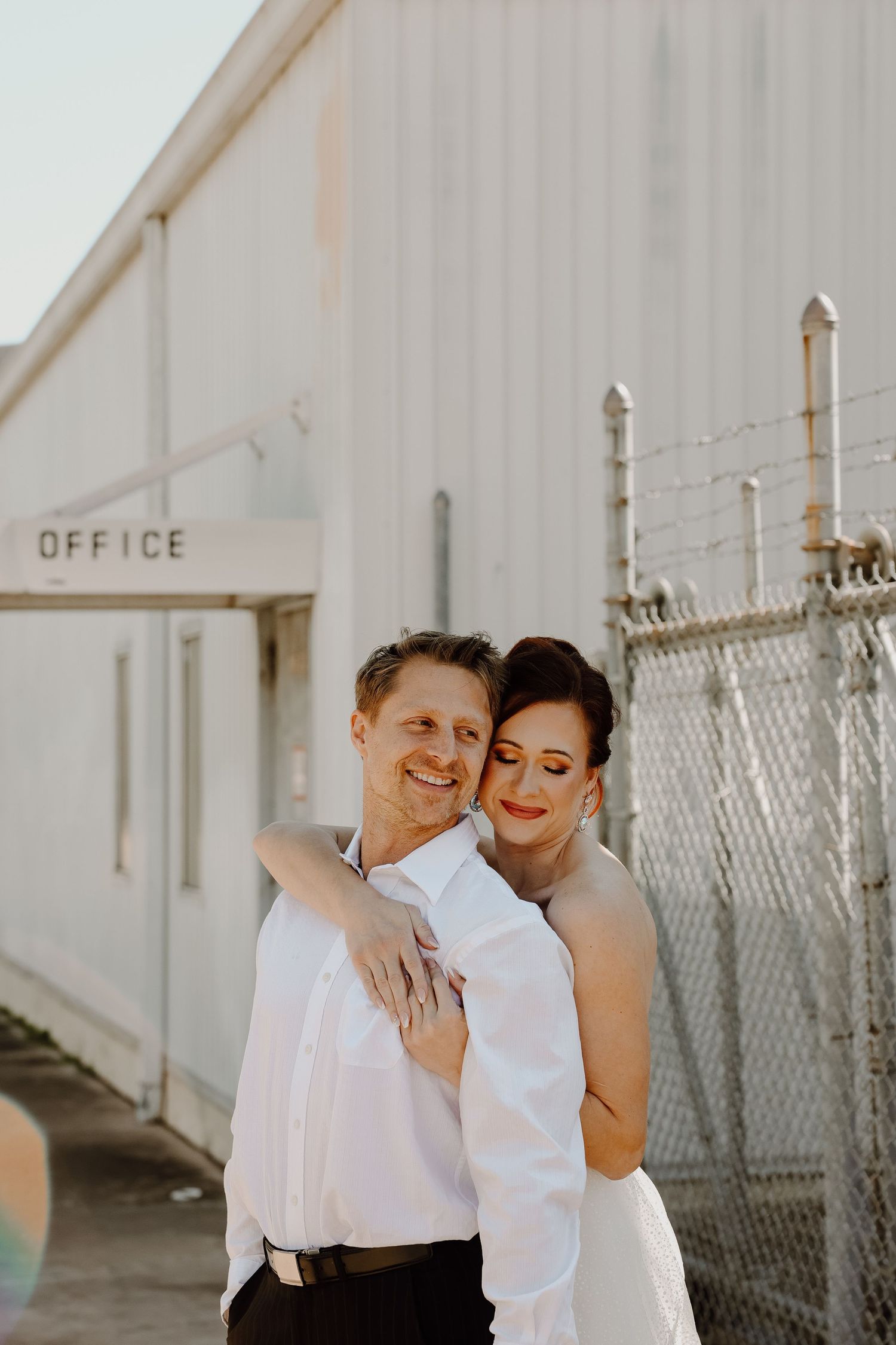 Smiling bride embracing groom from behind outside an office building with chain-link fence.
