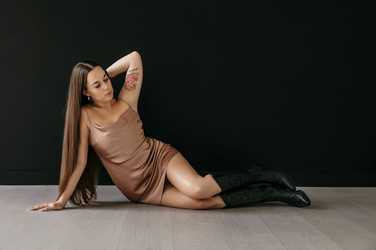 Woman in bronze satin slip dress posing on floor against dark wall with black knee-high boots.