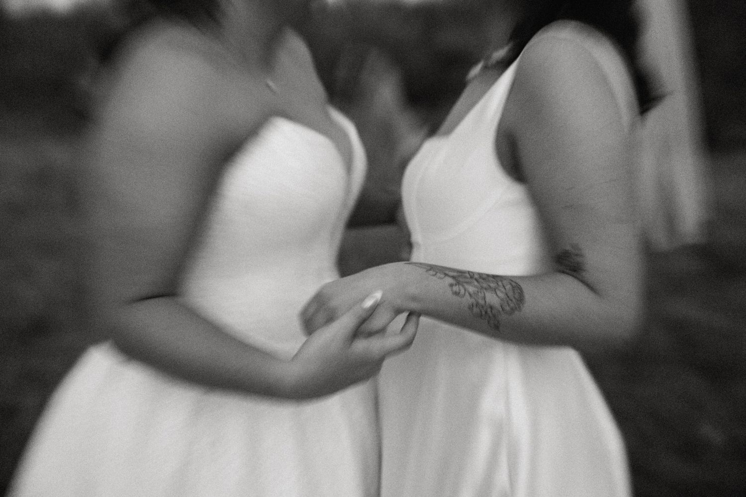 Artistic blurred black and white close-up of two brides holding hands, one with a floral tattoo.