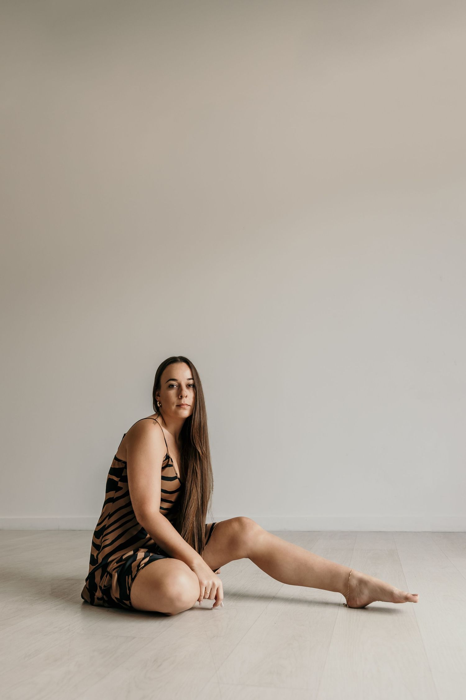 Woman in animal print slip dress sitting barefoot on floor against neutral wall in studio.