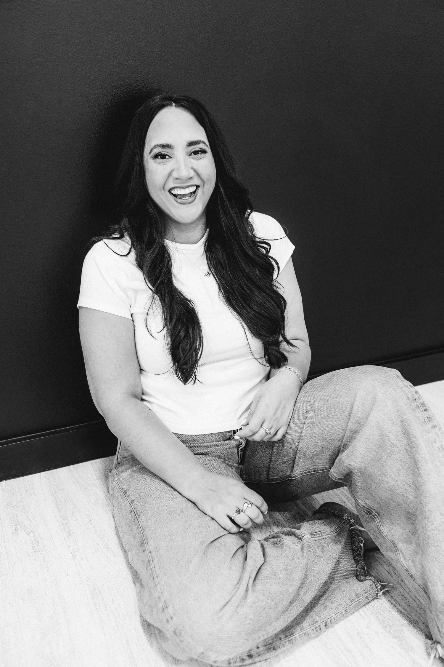 Woman in white tee and jeans laughing while sitting on floor, black and white photo.