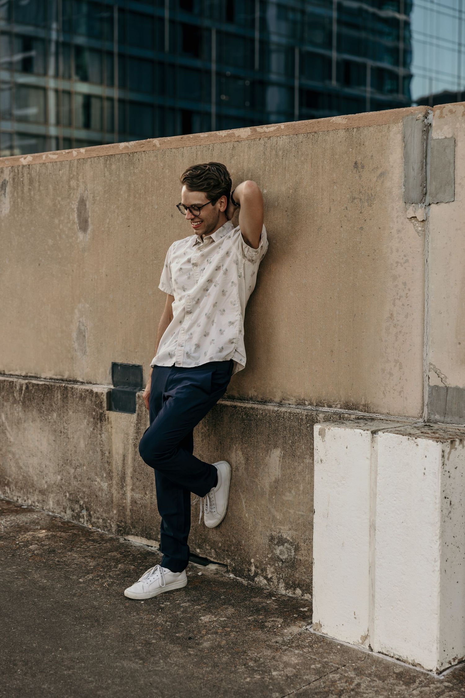 Stylish man in patterned shirt and dark pants leans against a concrete wall near city buildings.