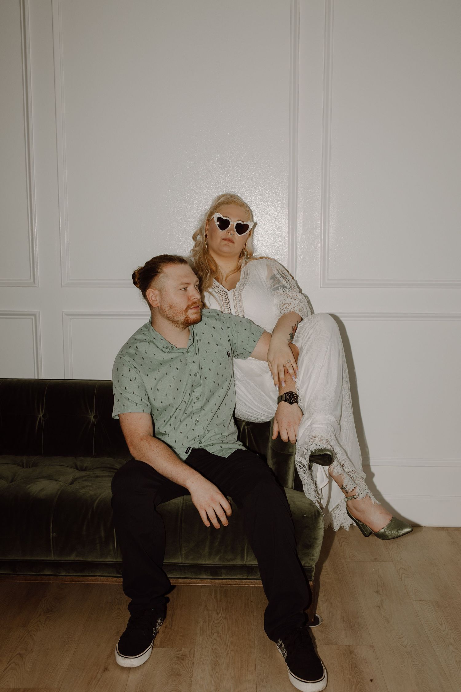 Stylish couple poses on a green velvet sofa, woman in white outfit with sunglasses resting on mans shoulder.