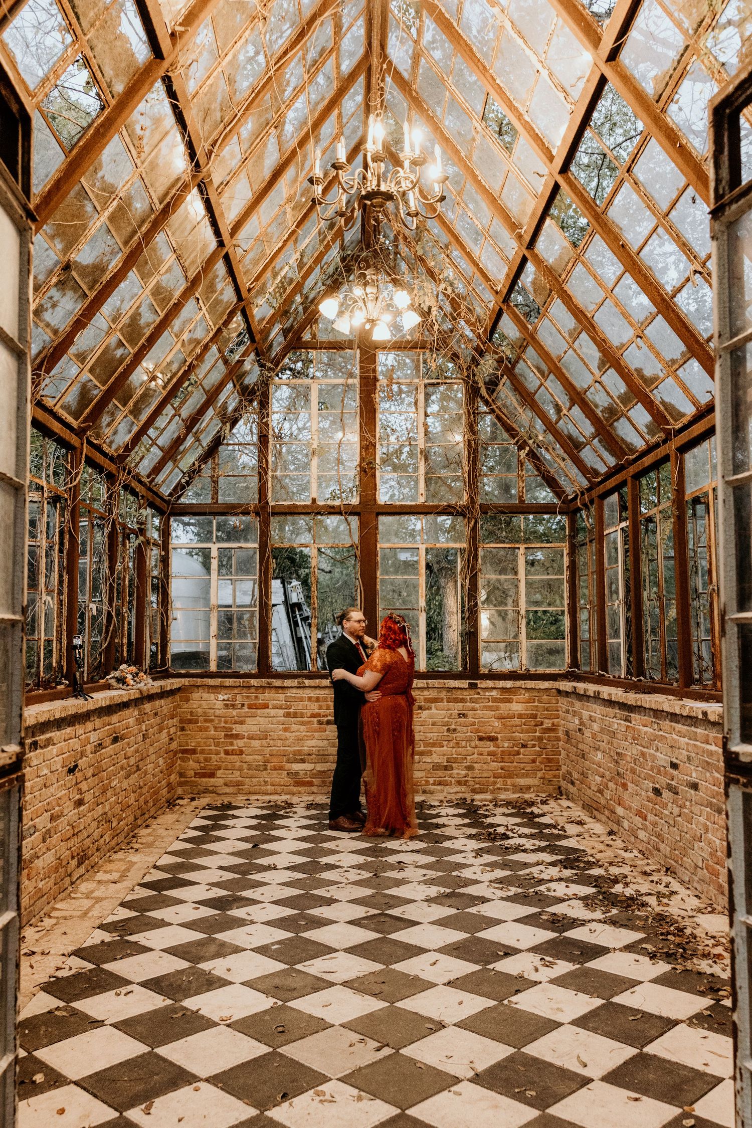 Couple shares romantic moment in rustic glass greenhouse with chandelier and checkered tile floor.