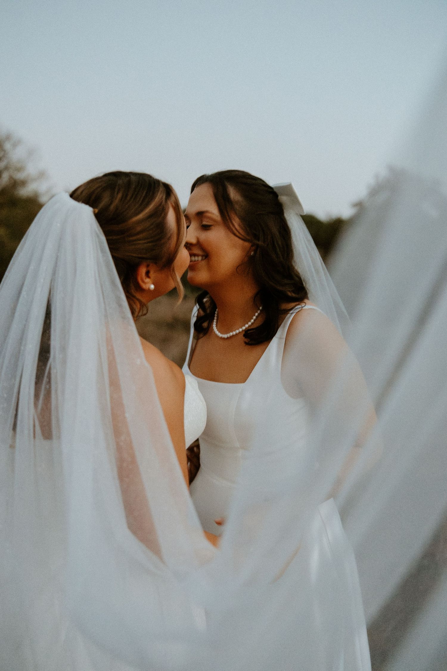 Two brides in white gowns and veils smiling face to face outdoors at dusk in romantic portrait.