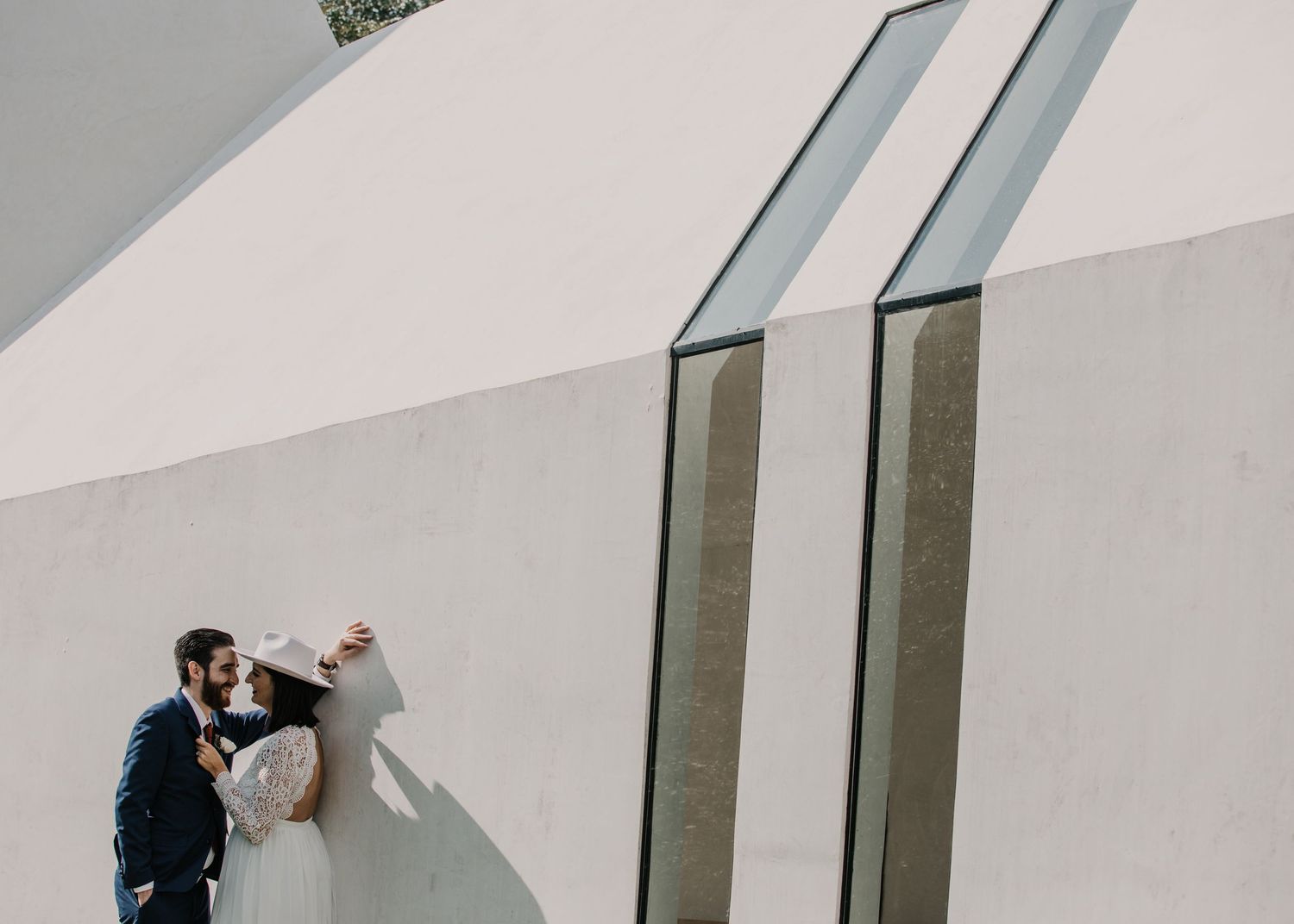 Aerial view of bride and groom sharing a kiss against a modern white architectural wall with geometric glass details.