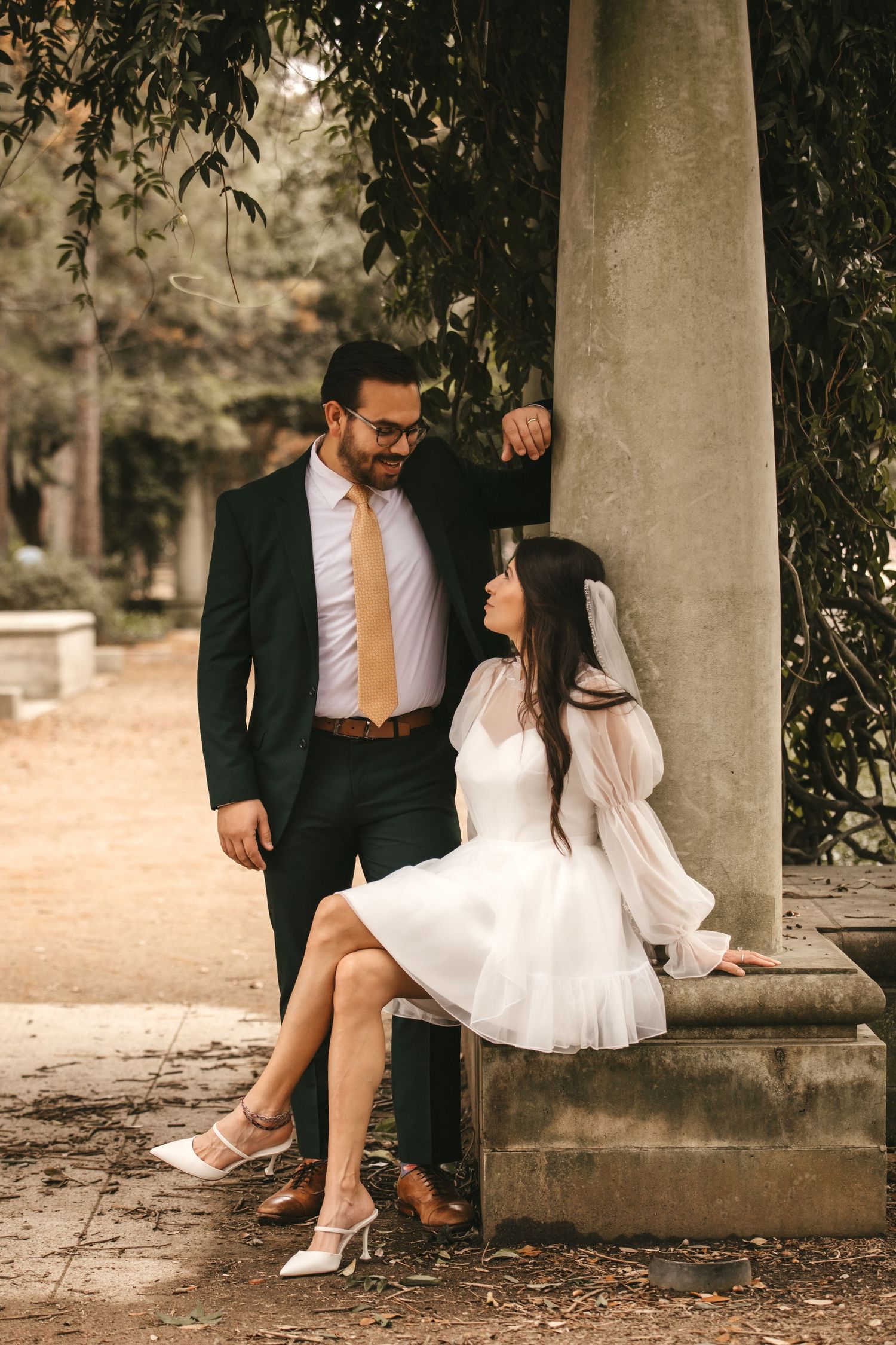 Bride in short white dress sits on stone pillar as groom in dark suit leans over her in a park setting.