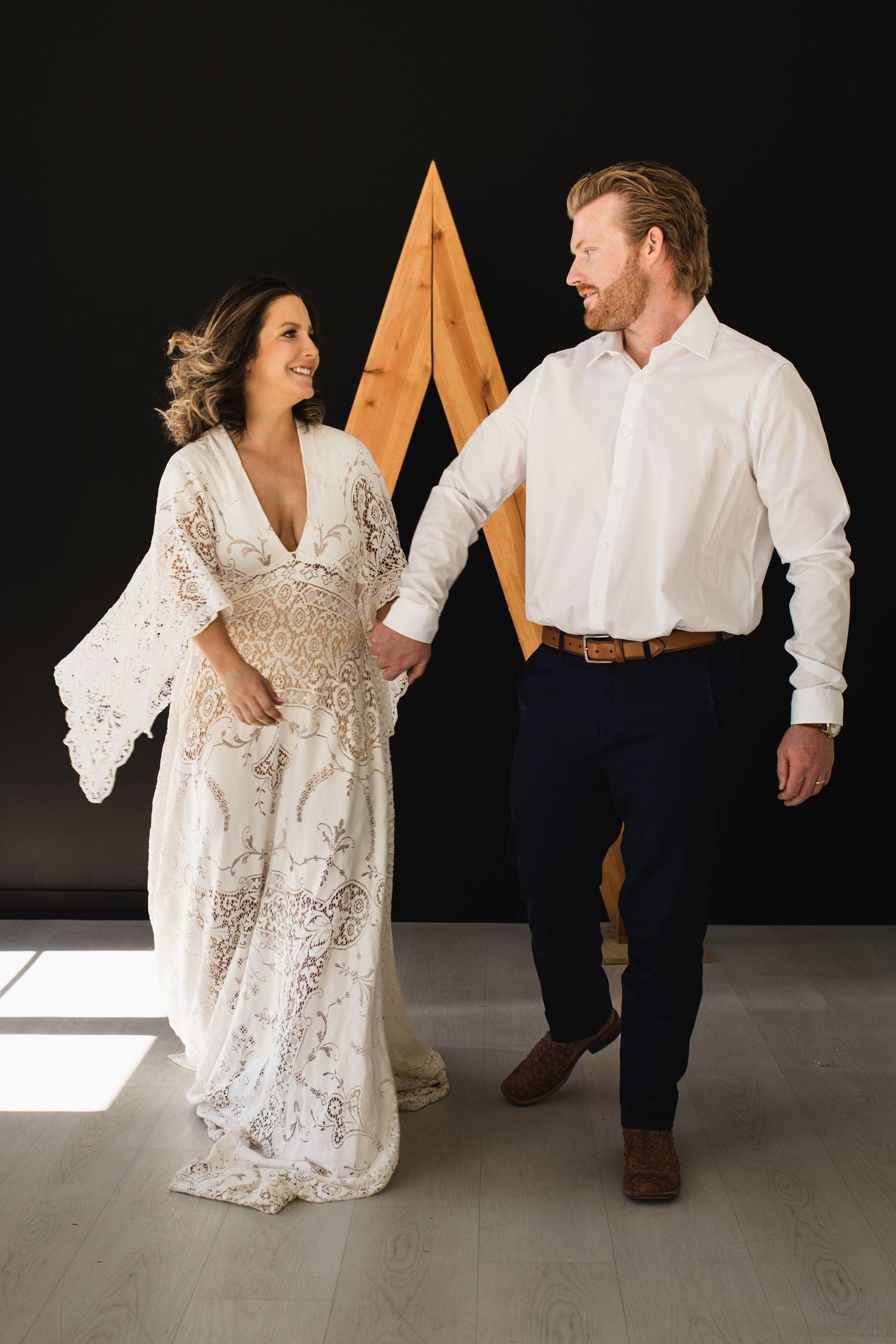 Couple in bohemian wedding attire holding hands, smiling, with a wooden geometric backdrop.