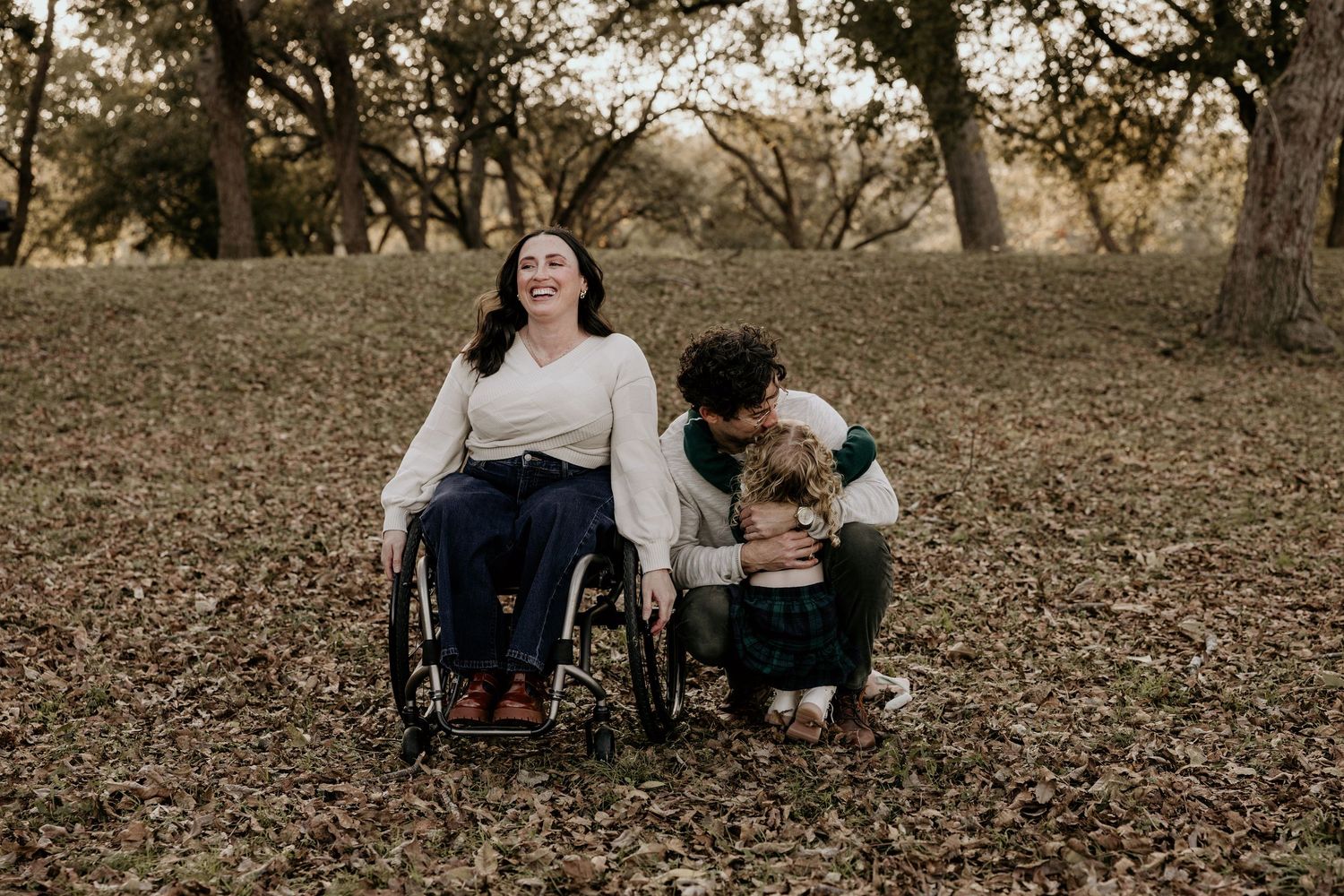 Woman in wheelchair laughing with man crouching beside her in a leaf-covered autumn park setting.