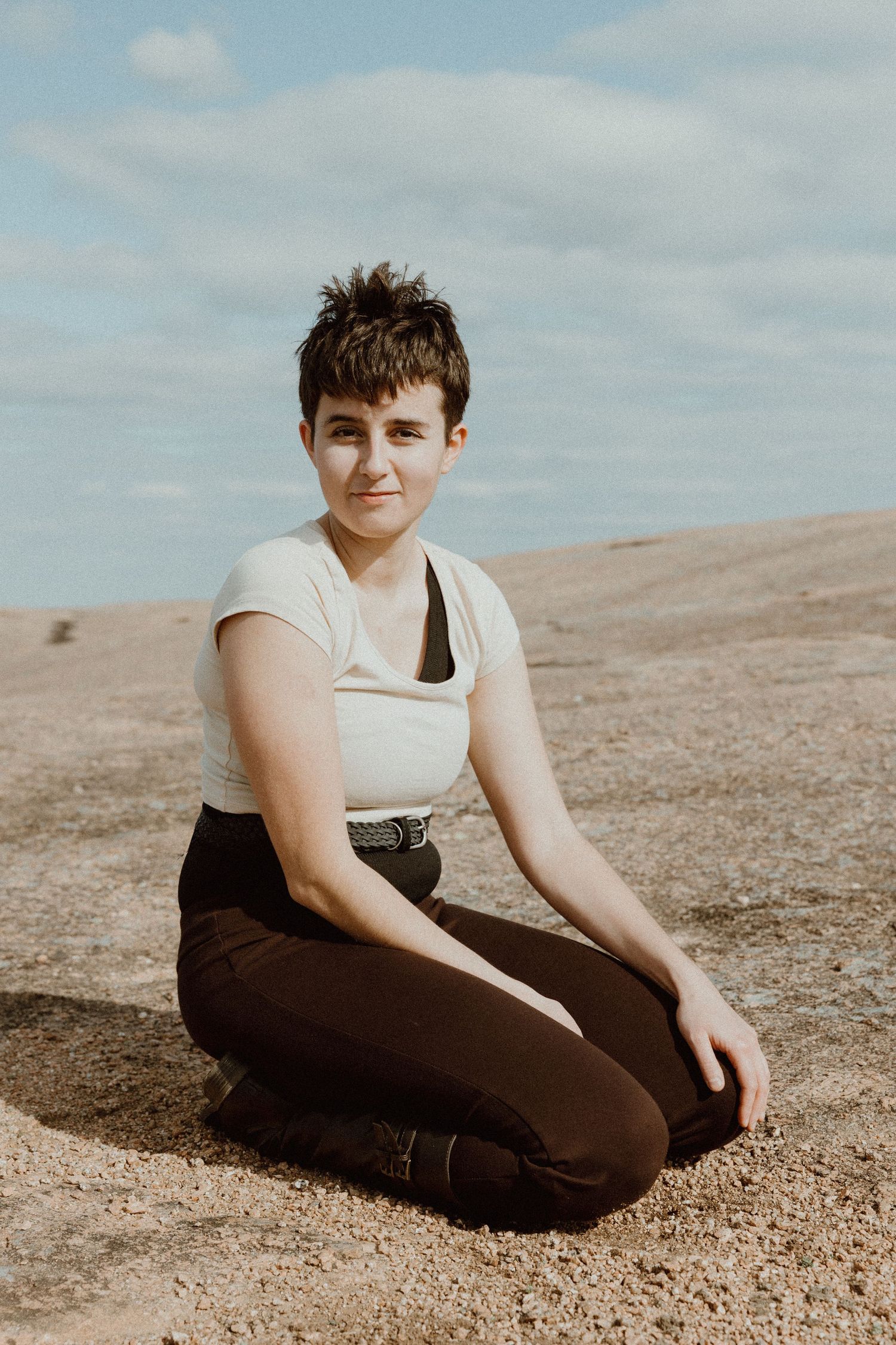 Short-haired person in white top and dark pants sitting cross-legged on rocky ground.