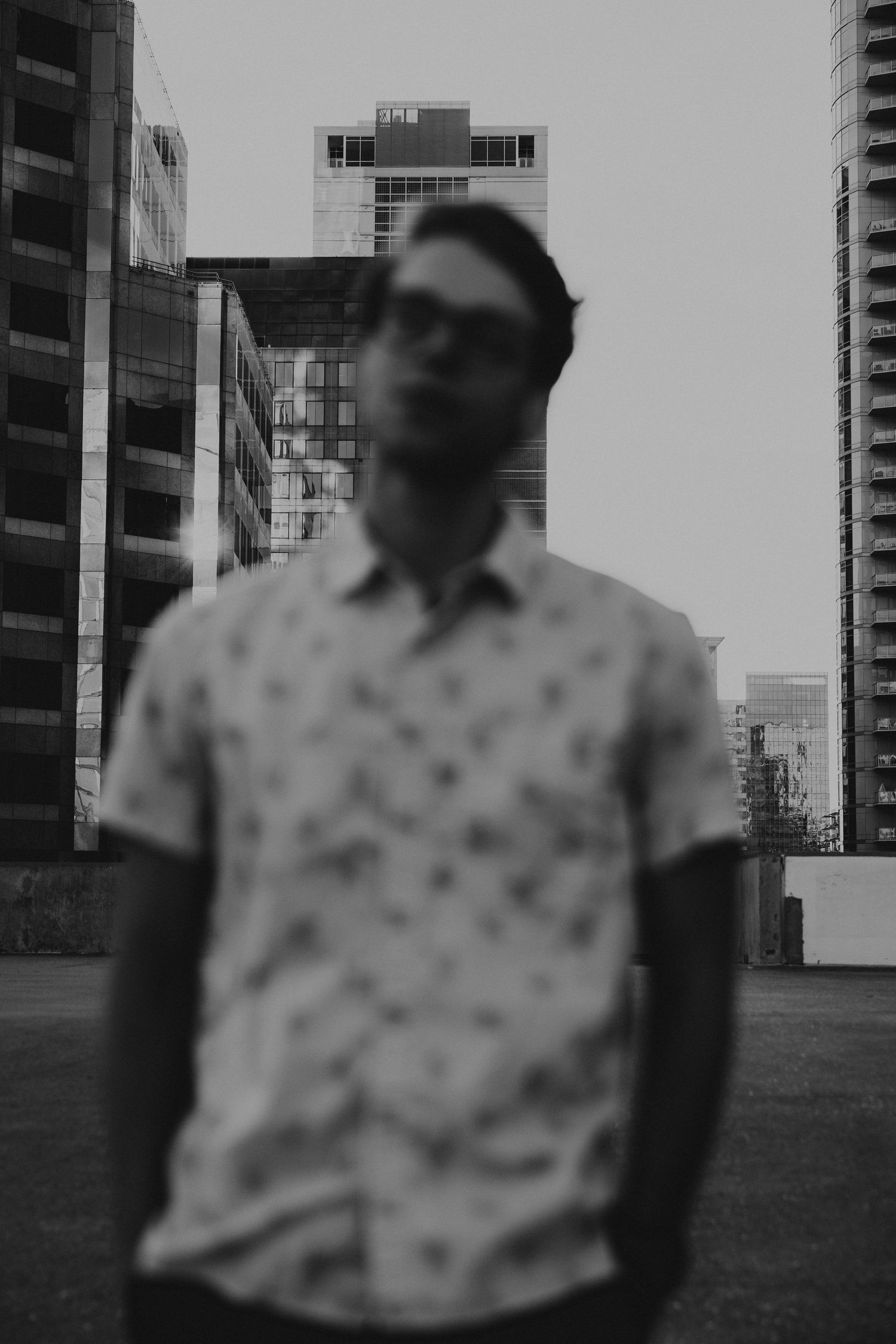 Artistic blurred black and white portrait of a man in a patterned shirt with city buildings behind.