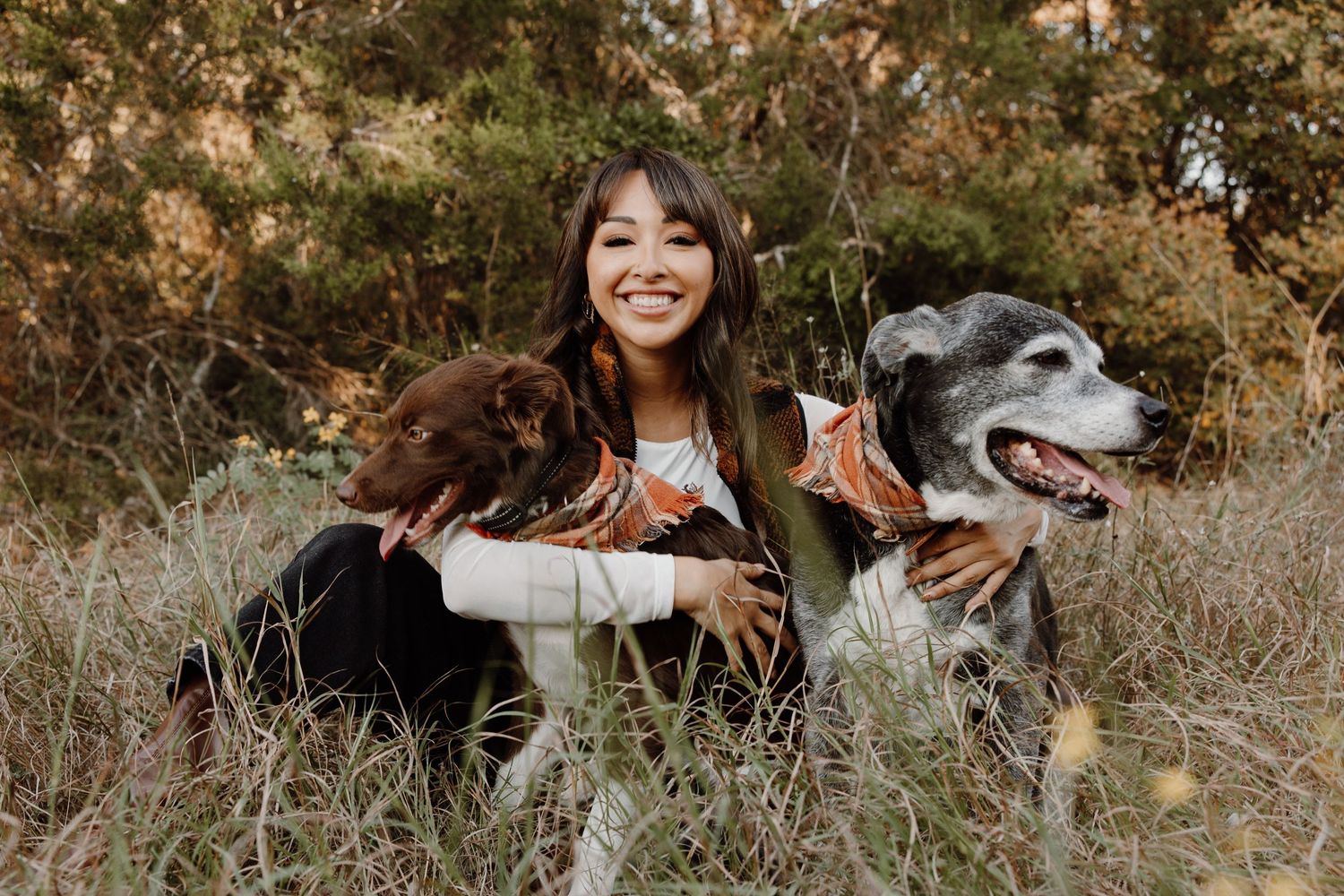 Smiling young woman sitting in tall grass with two happy dogs in a natural outdoor setting.