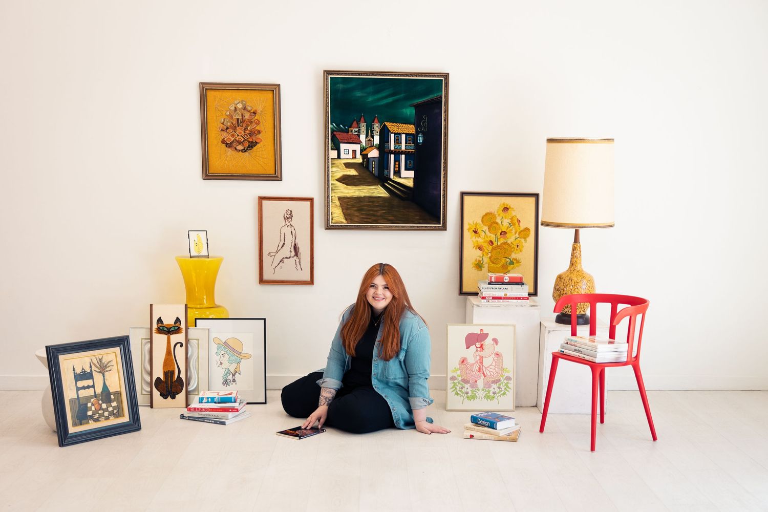 Redheaded woman sitting on floor surrounded by colorful vintage paintings and artwork in white room.