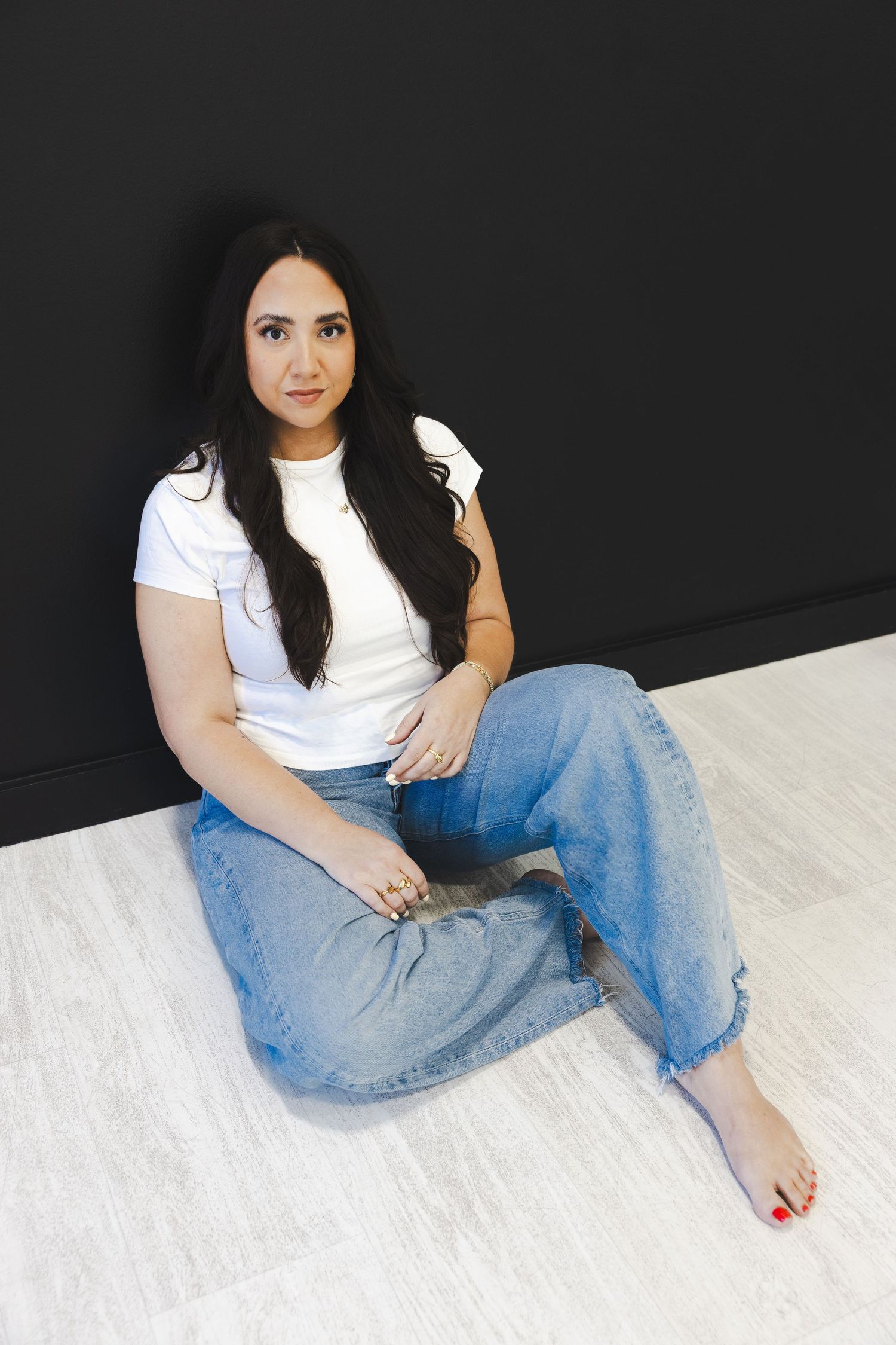 Woman in white tee and jeans sitting barefoot on floor against black wall.