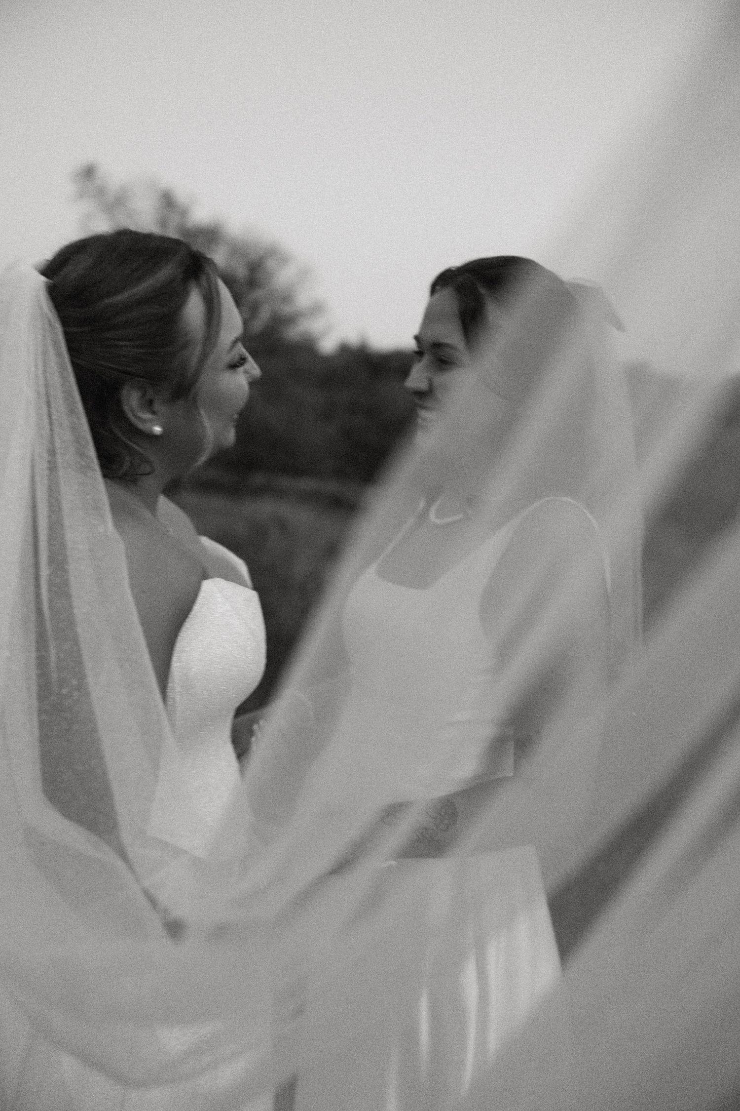 Two brides in white wedding gowns and veils face each other tenderly in a black and white portrait.