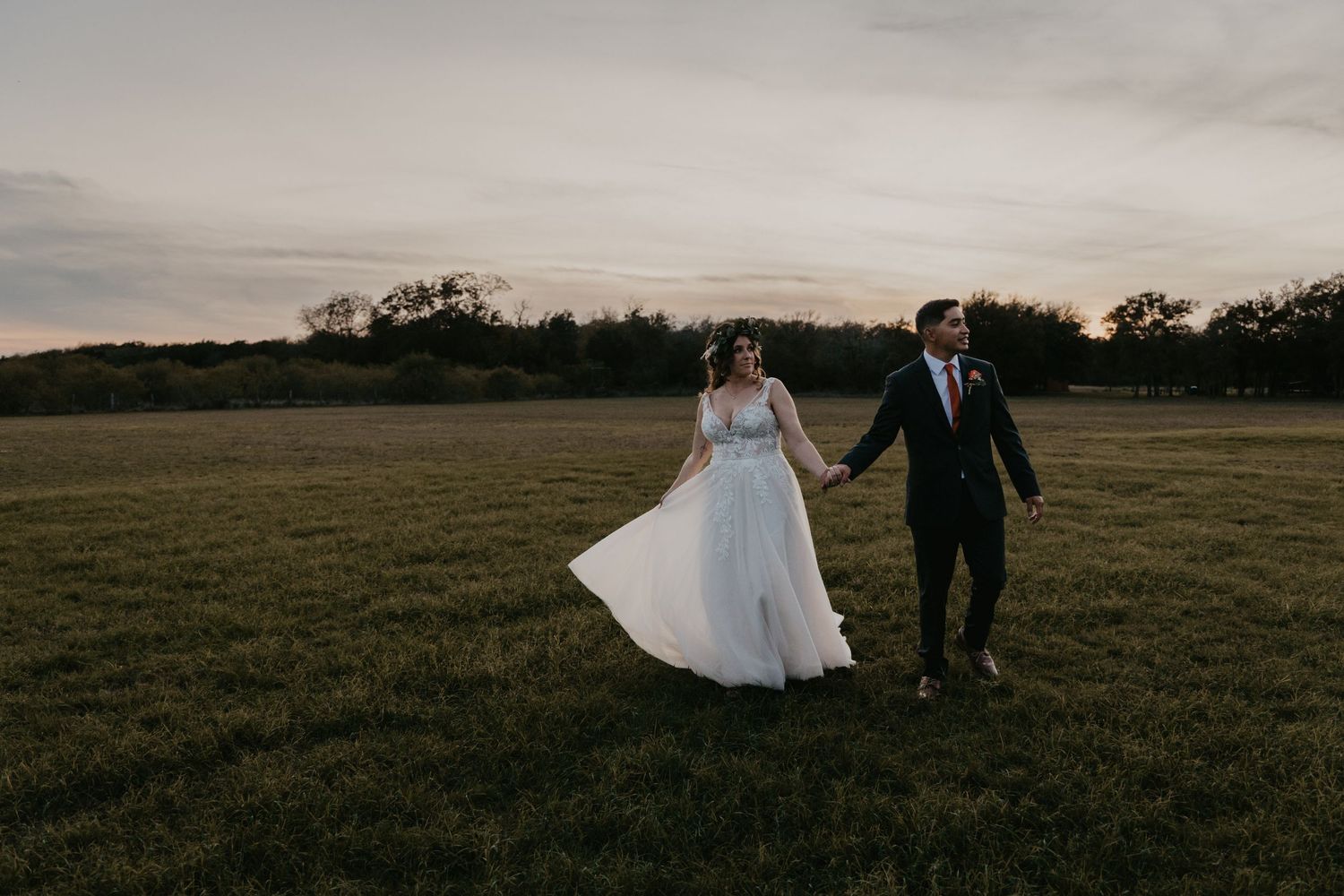 Bride and groom walk hand in hand across a vast green field at golden hour sunset.