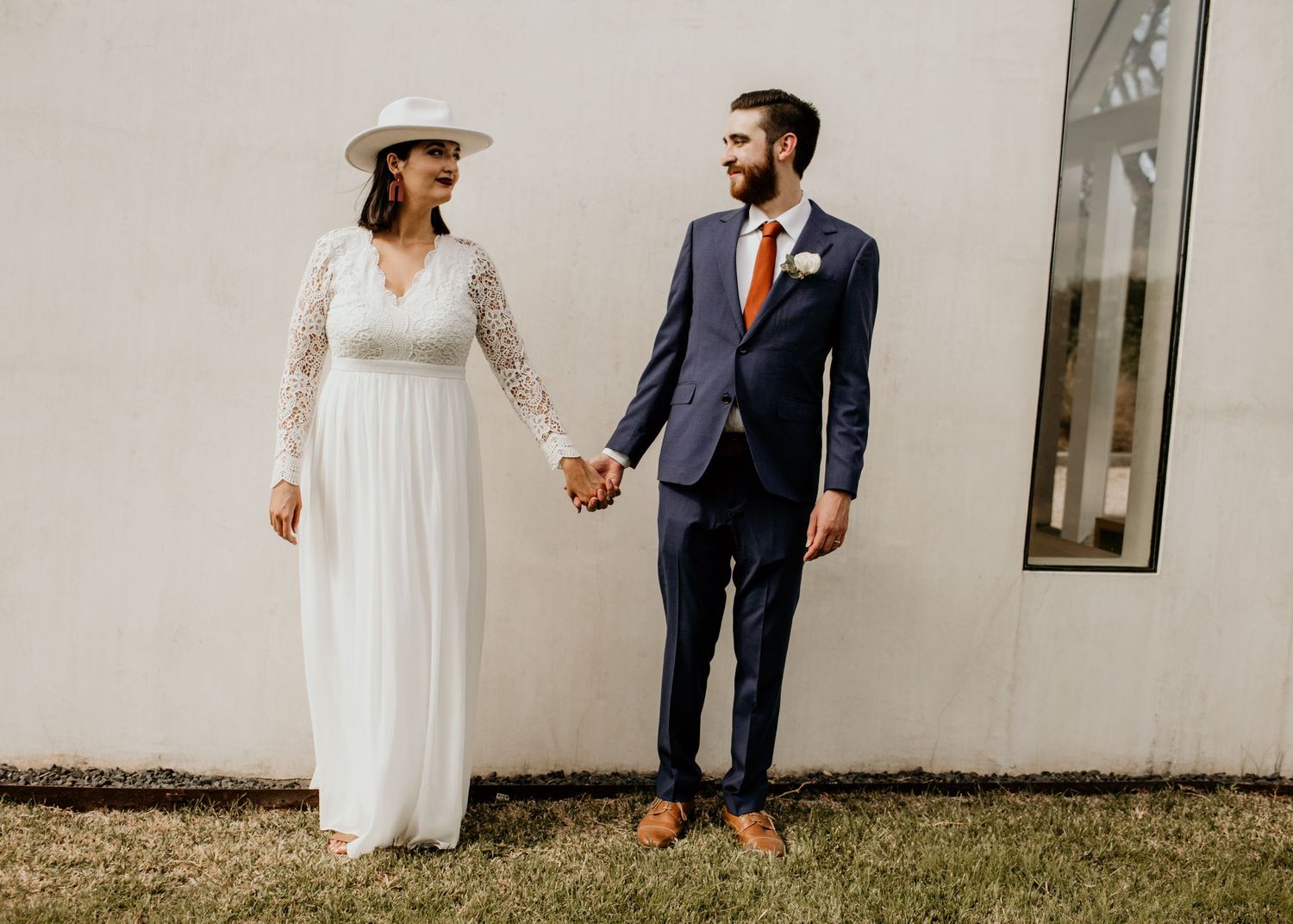 Bride in white lace gown and hat holds hands with groom in navy suit outdoors at wedding venue.