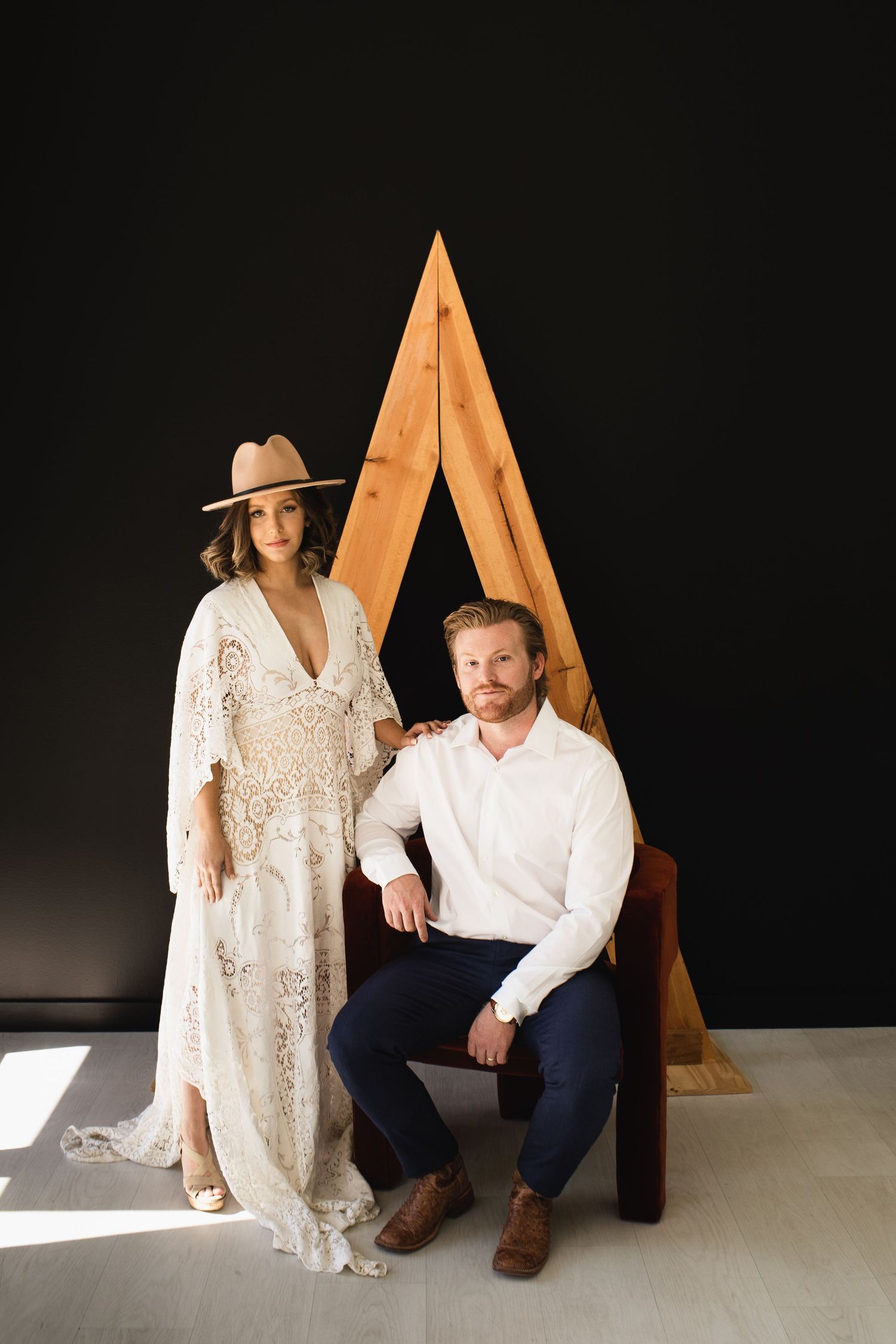 Boho bride in lace dress and hat poses with groom in white shirt before a wooden triangle backdrop.