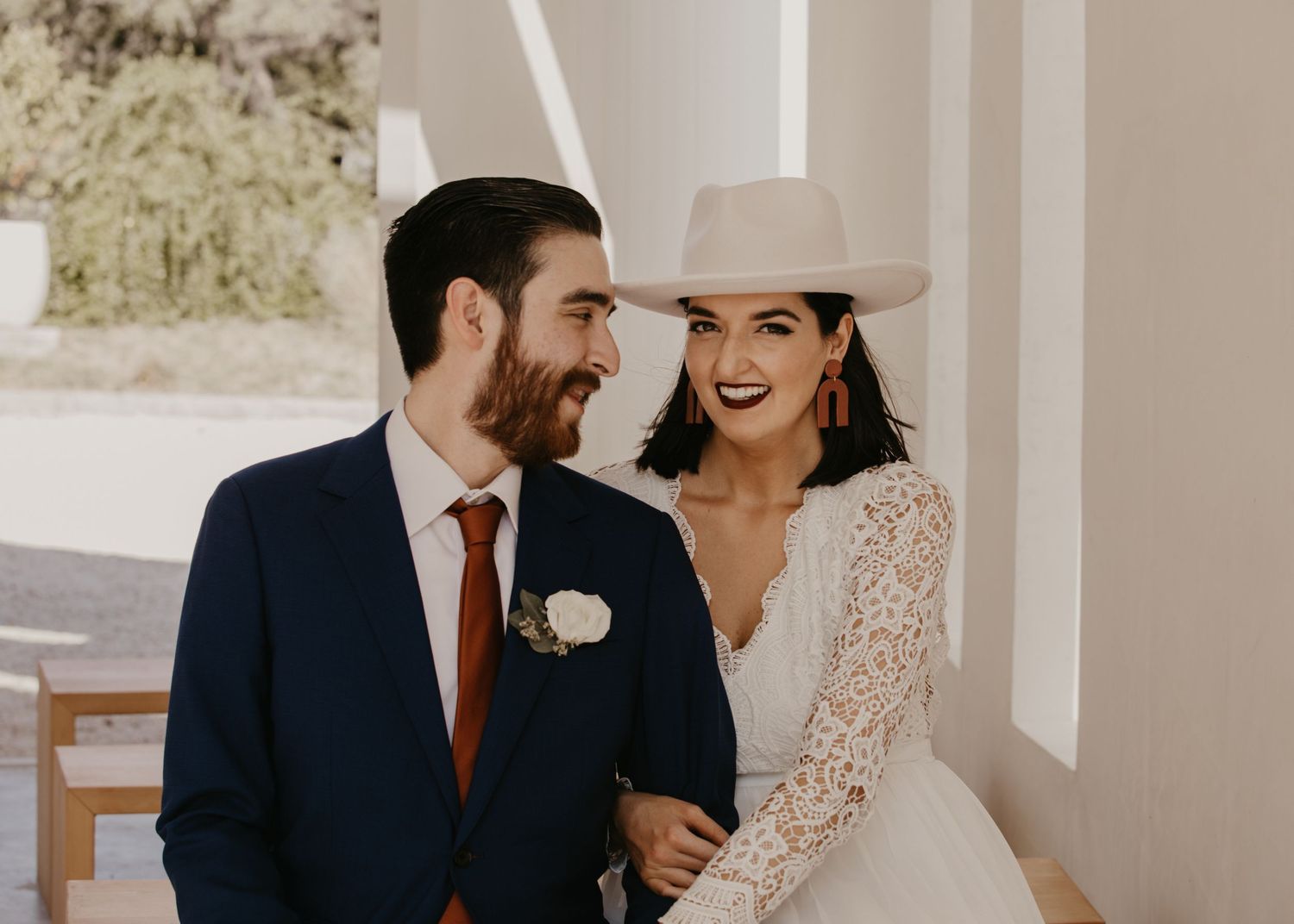 Groom in navy suit with rust tie smiling at bride wearing a white cowboy hat and lace wedding dress.
