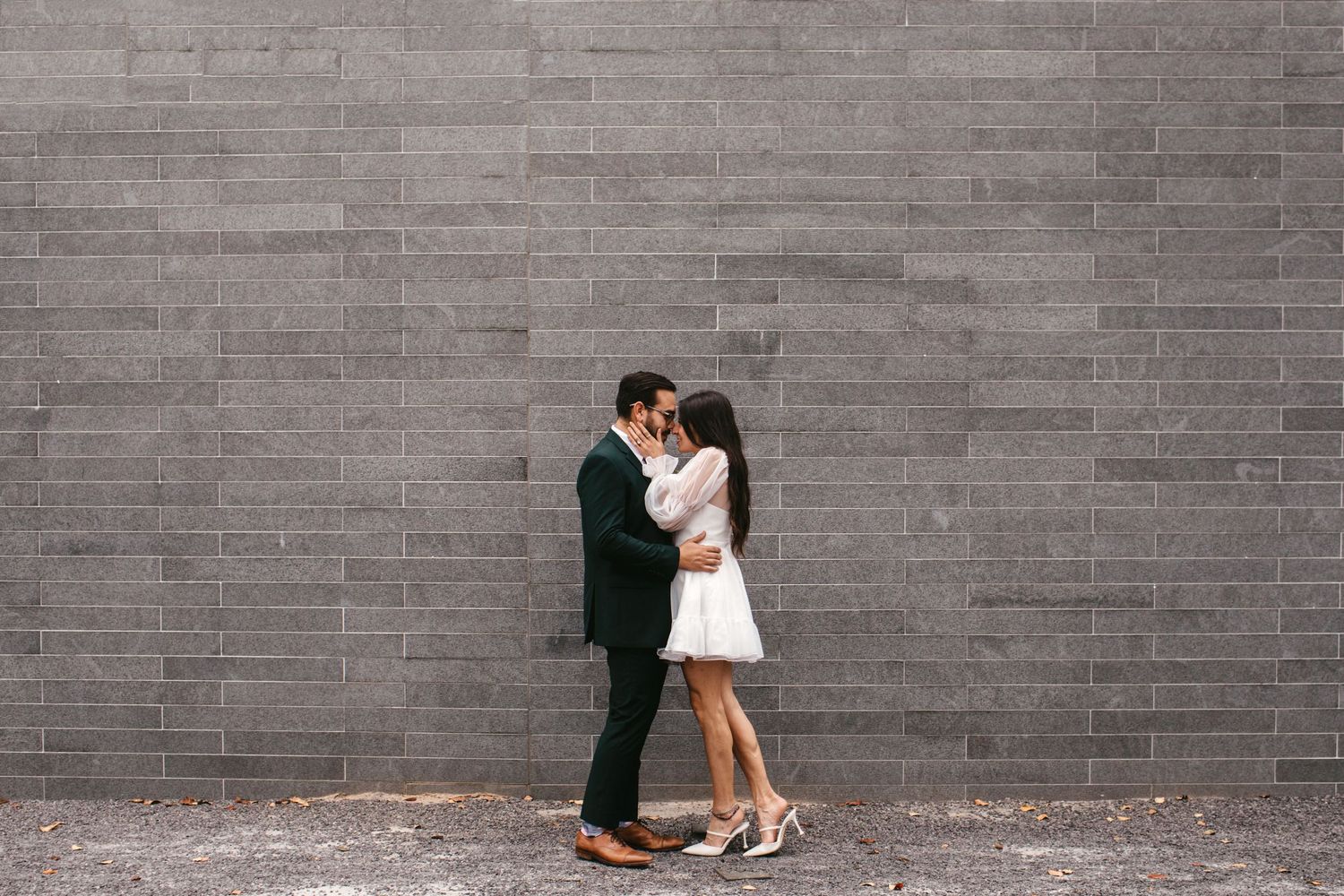 Bride and groom share a kiss against a gray brick wall, she in a white mini dress and he in a dark suit.