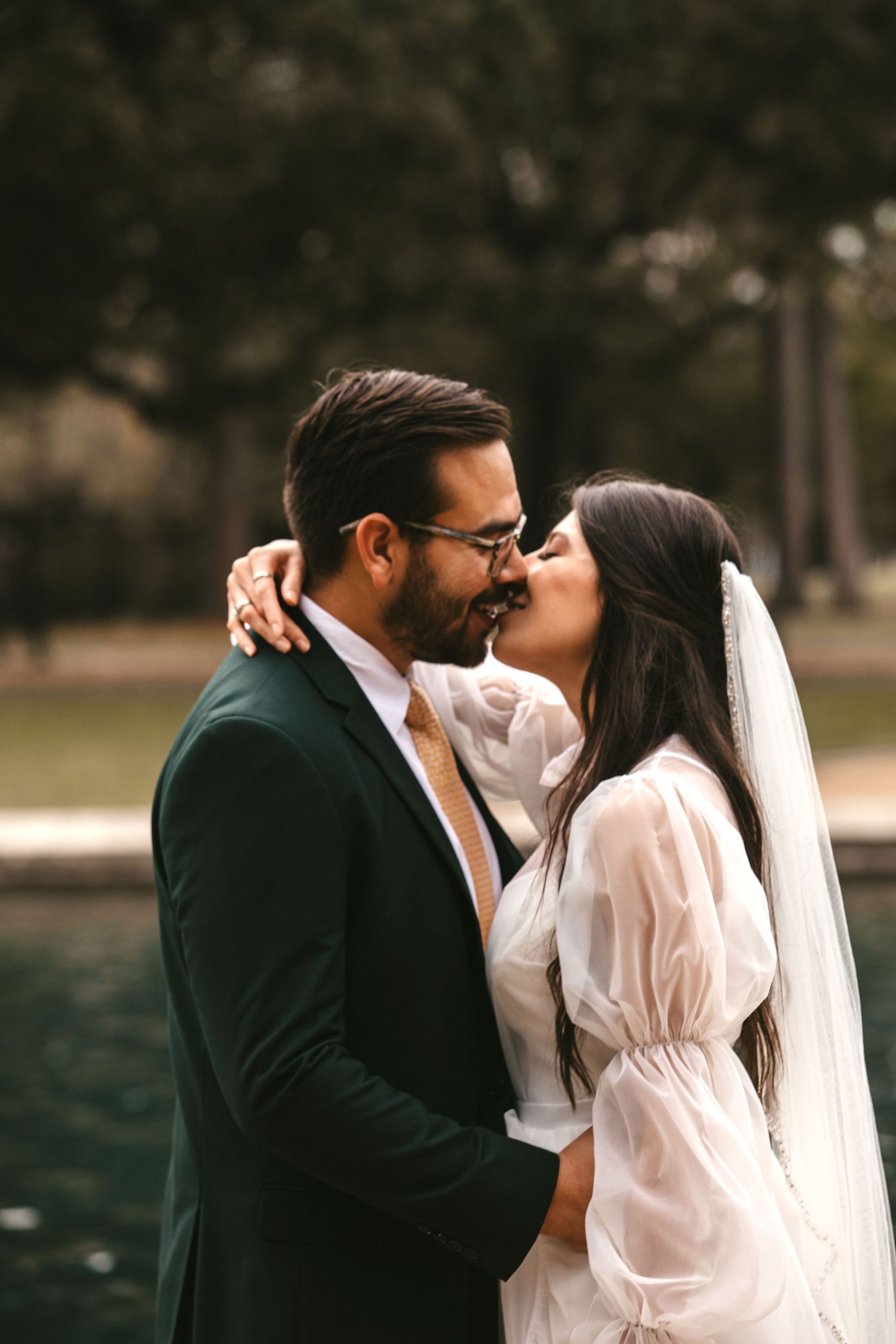 Bride and groom share a kiss near a pond surrounded by lush green trees.