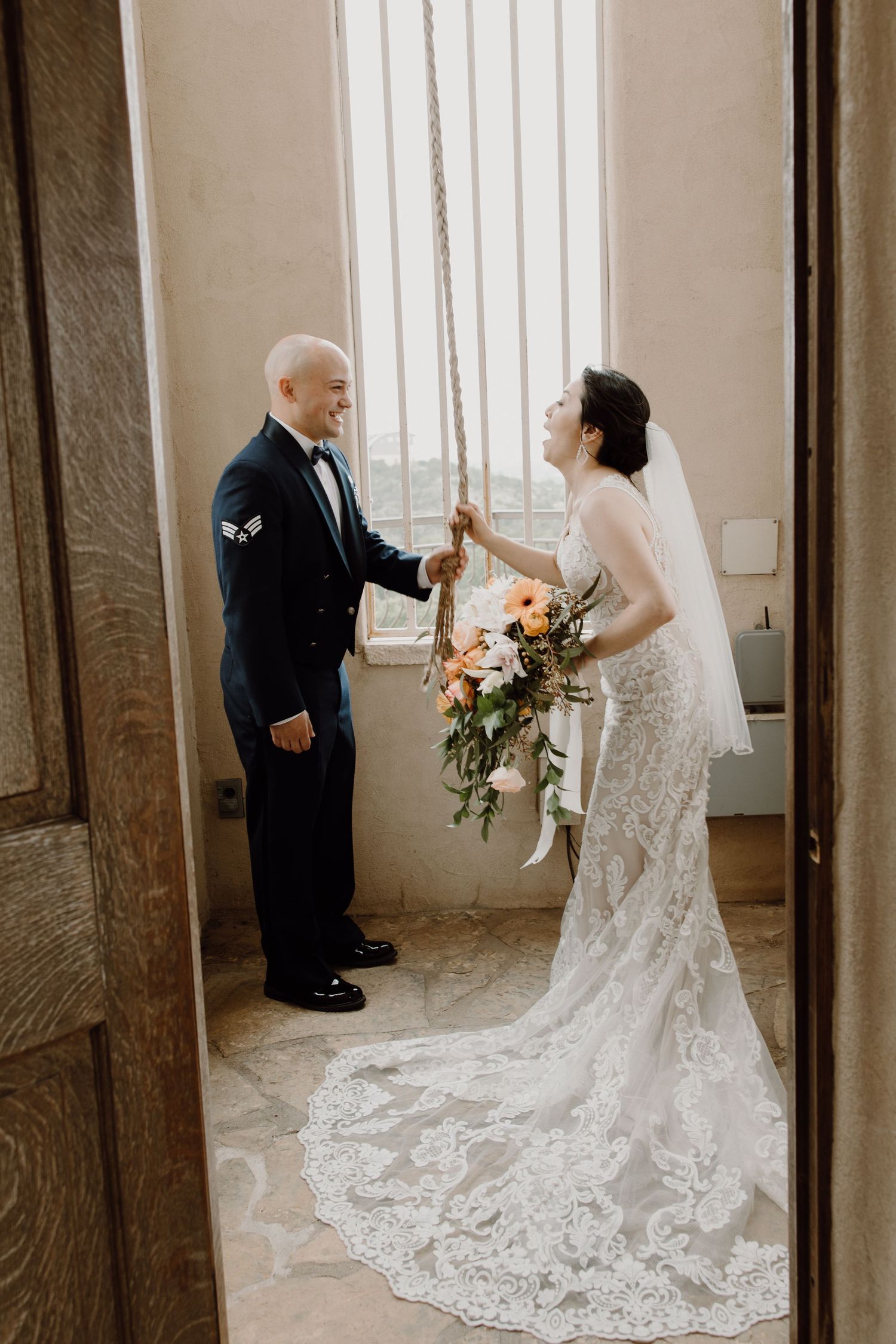 Bride in lace gown holds colorful bouquet while sharing a tender moment with groom in military uniform indoors.
