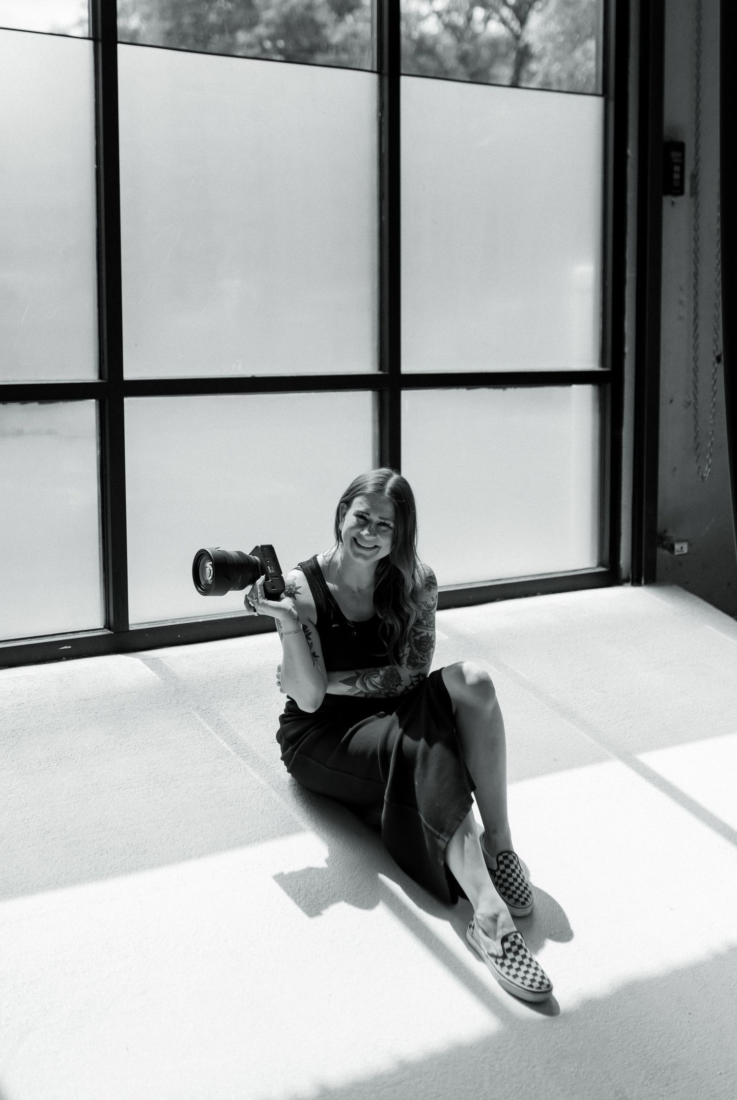 Black and white photo of tattooed photographer sitting on floor holding camera by windows.