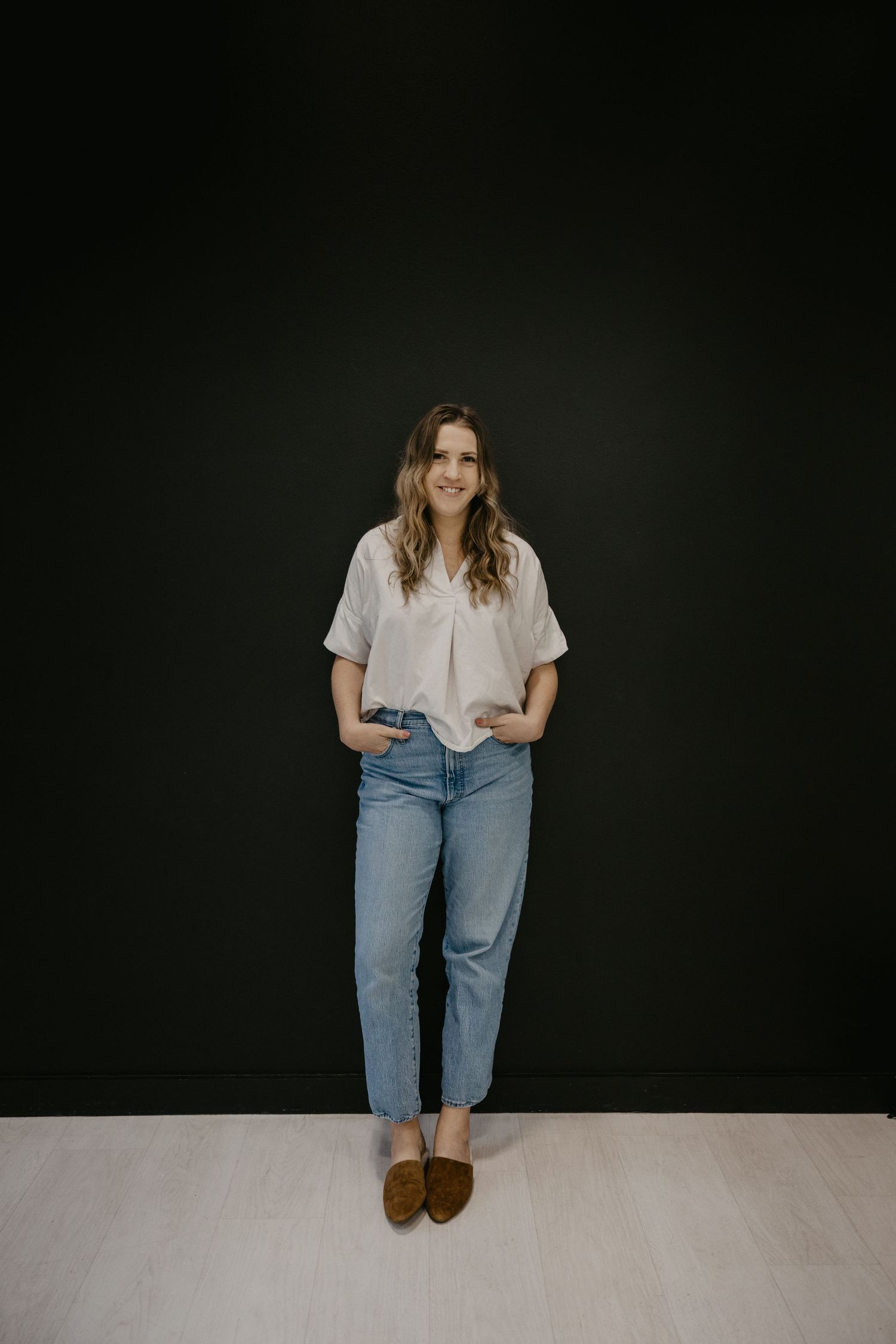 Full length portrait of a woman in white top and jeans standing against a dark wall.