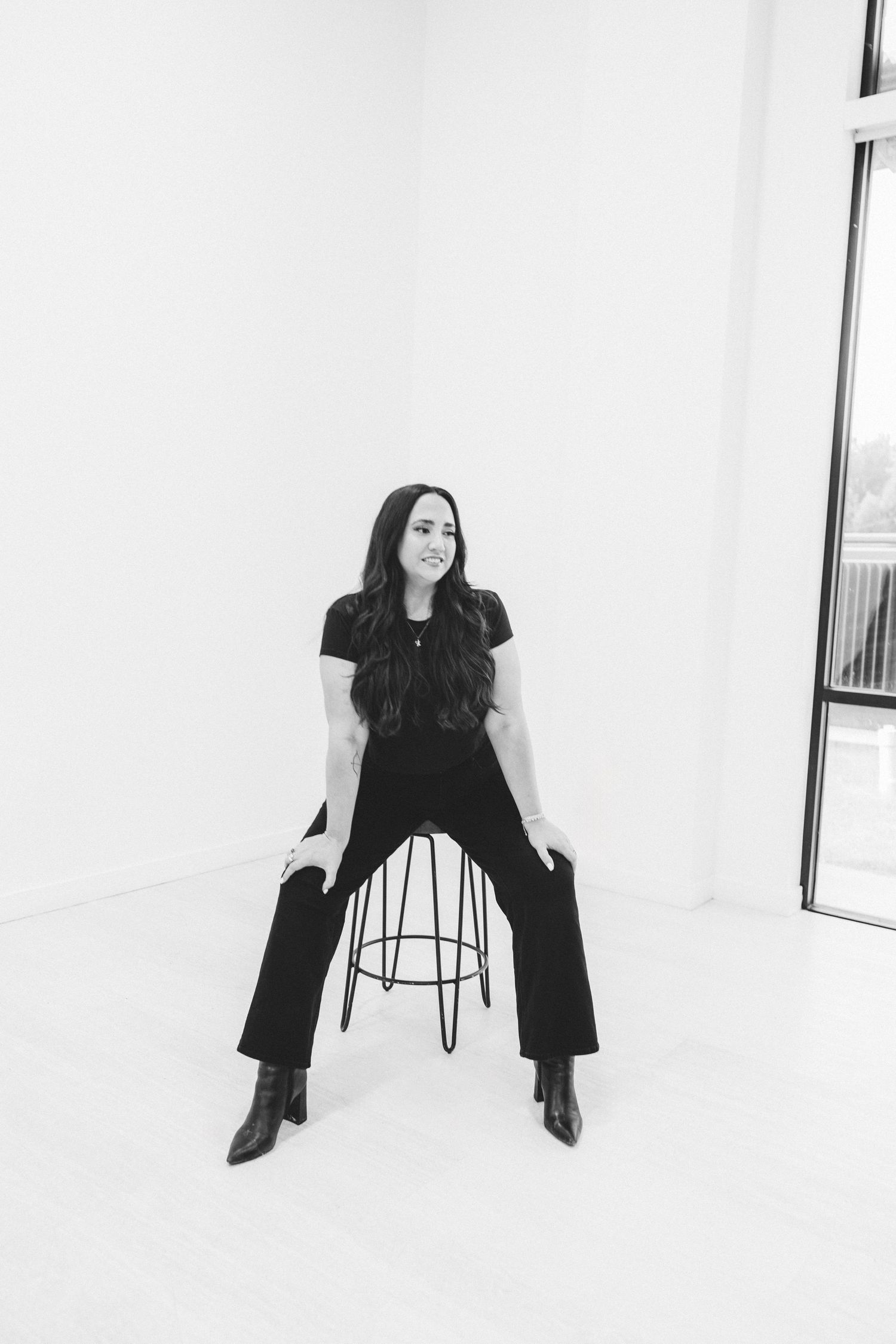 Black and white portrait of a woman sitting on a stool in a bright minimalist white studio.