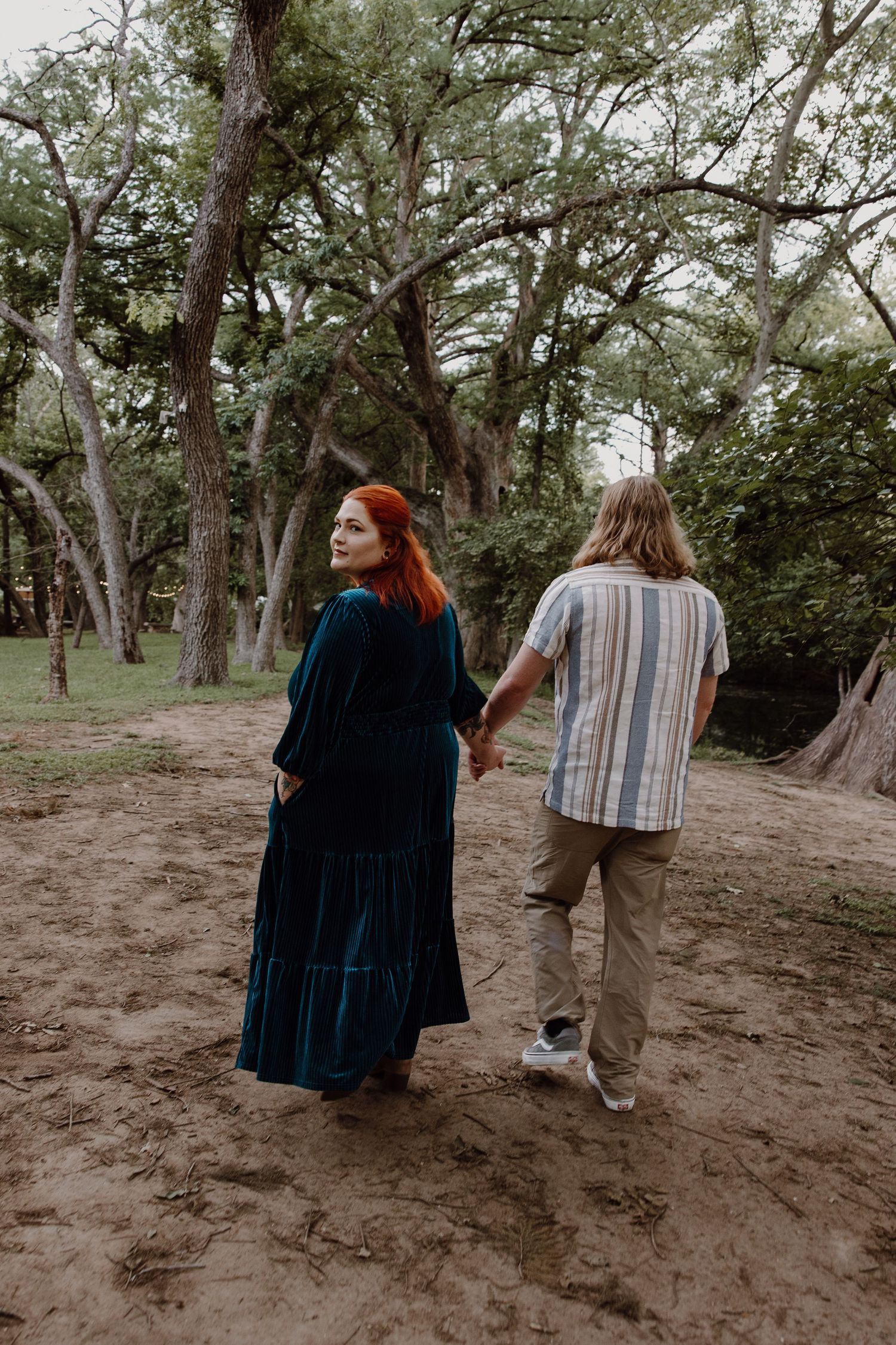 A woman in a teal velvet maxi dress holds hands with a man in a striped shirt as they walk through a sandy forest.