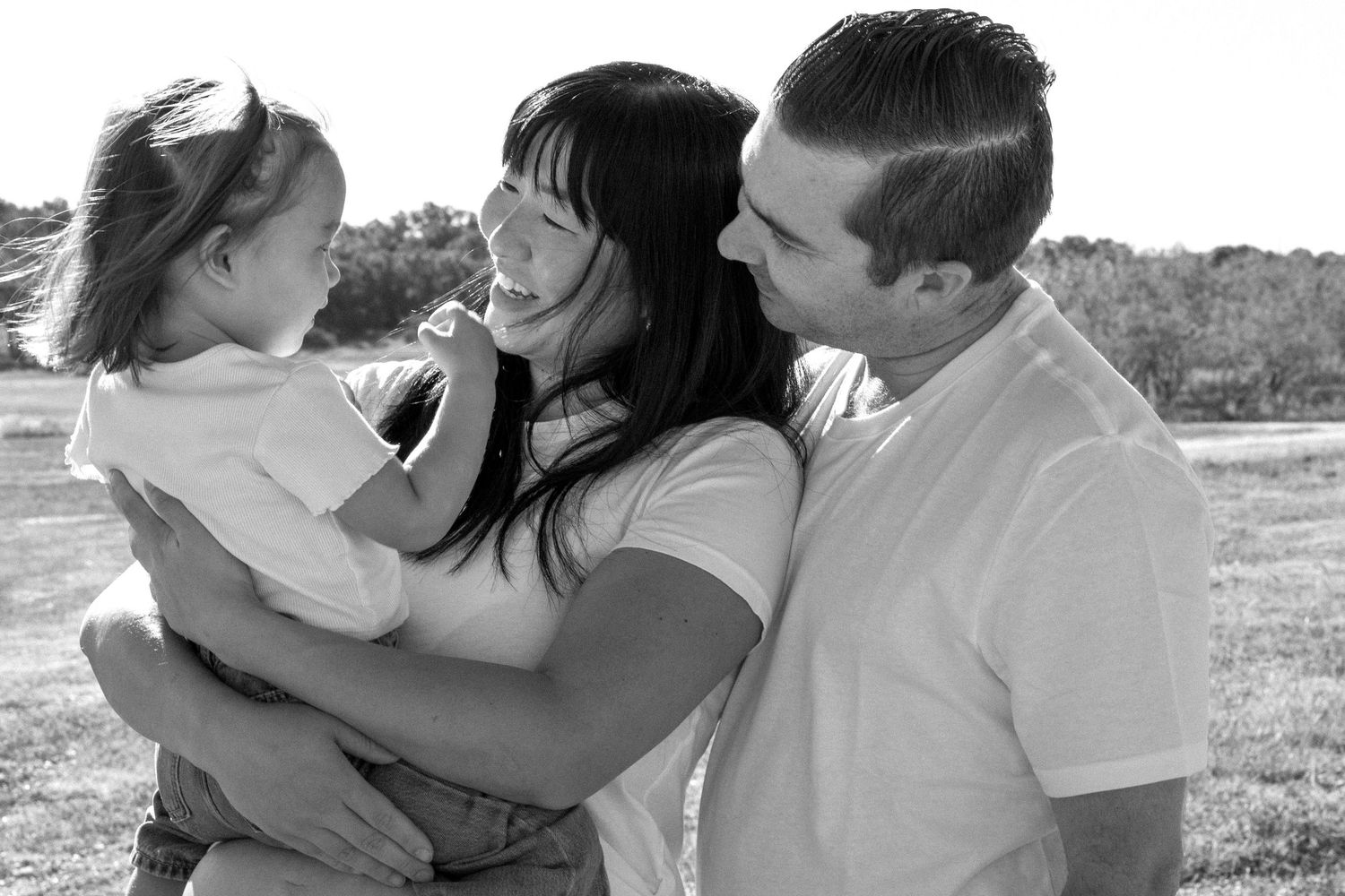 Black and white photo of a happy family with mother, father, and young daughter sharing a joyful moment outdoors.