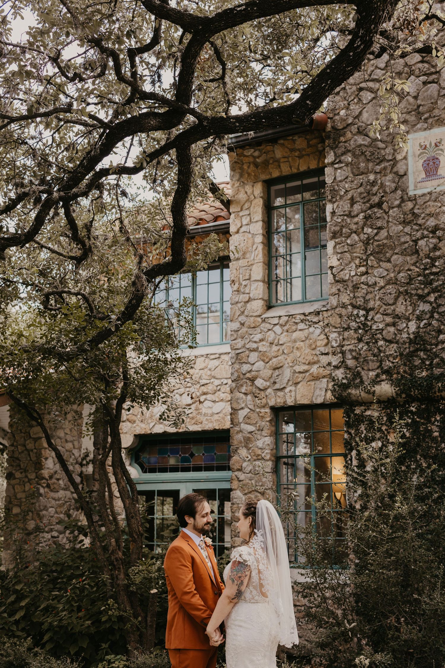 Bride and groom hold hands before a rustic stone building surrounded by oak trees at their wedding.