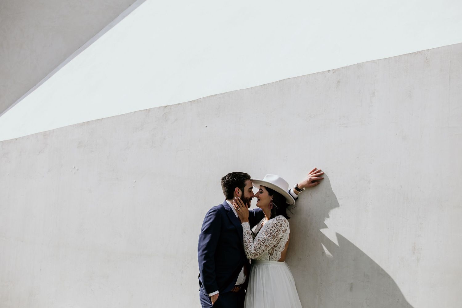 Couple sharing a romantic kiss against a white textured wall, bride in lace gown and sun hat, groom in navy suit.