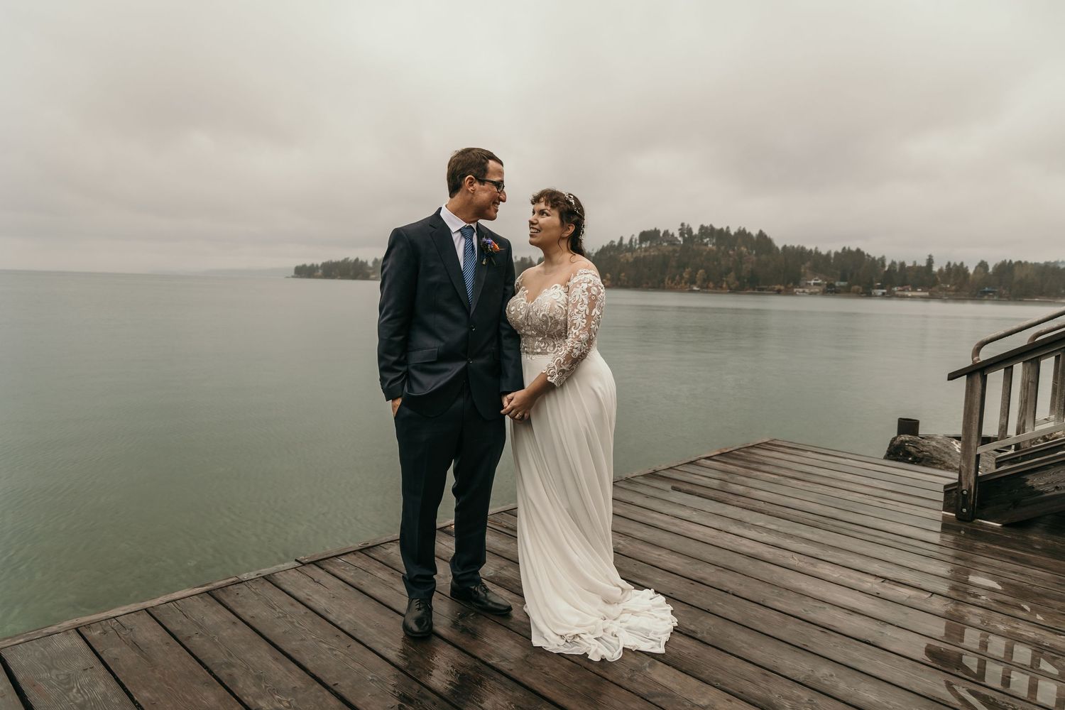 Bride in lace gown and groom in navy suit sharing a tender moment on a misty waterfront dock.