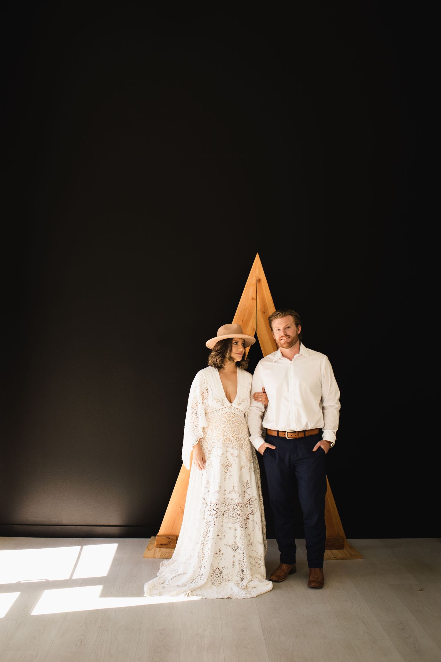 Boho bride in lace gown and wide-brim hat stands with groom before dark wall with wooden triangle backdrop.