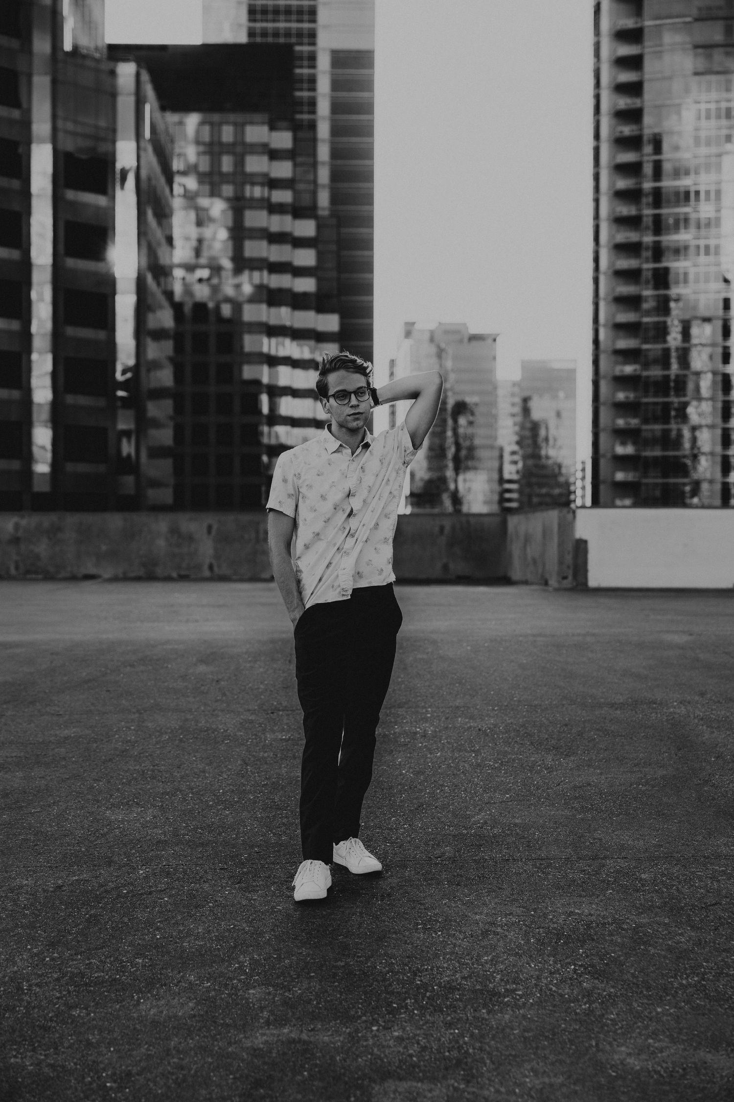 Black and white portrait of a man in a patterned shirt standing on a rooftop with city skyline.
