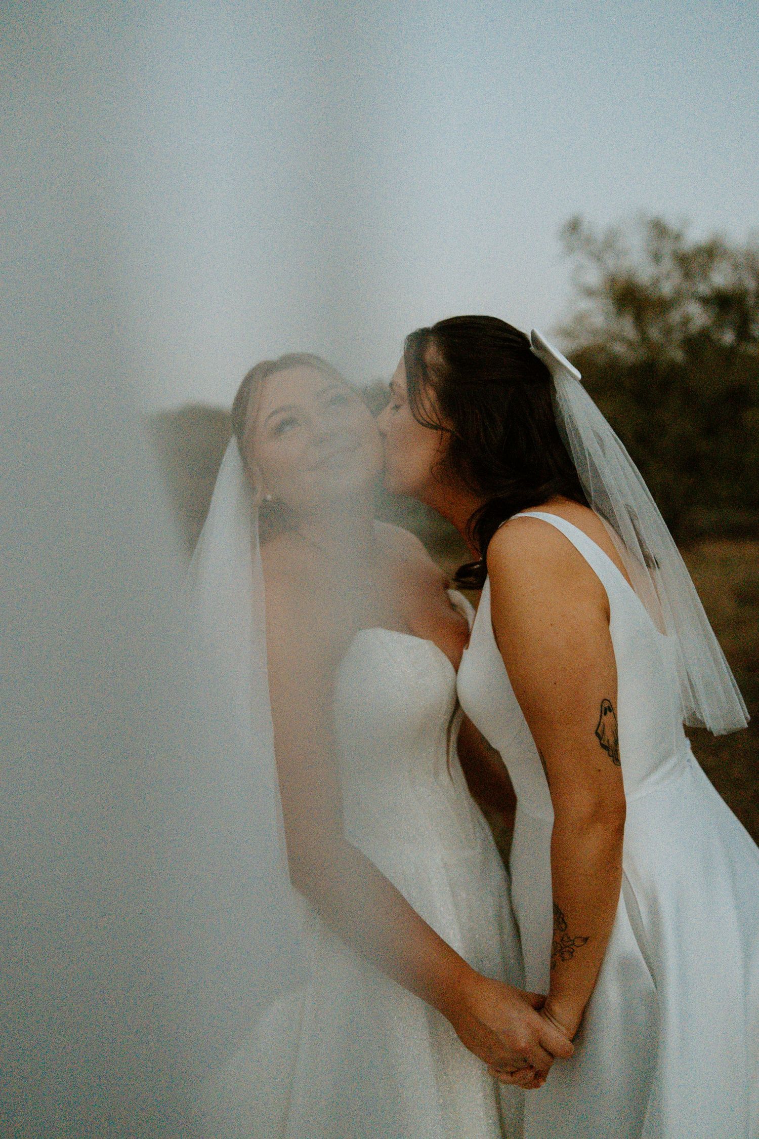 Two brides in white wedding dresses kissing tenderly with veil draped softly in foreground.