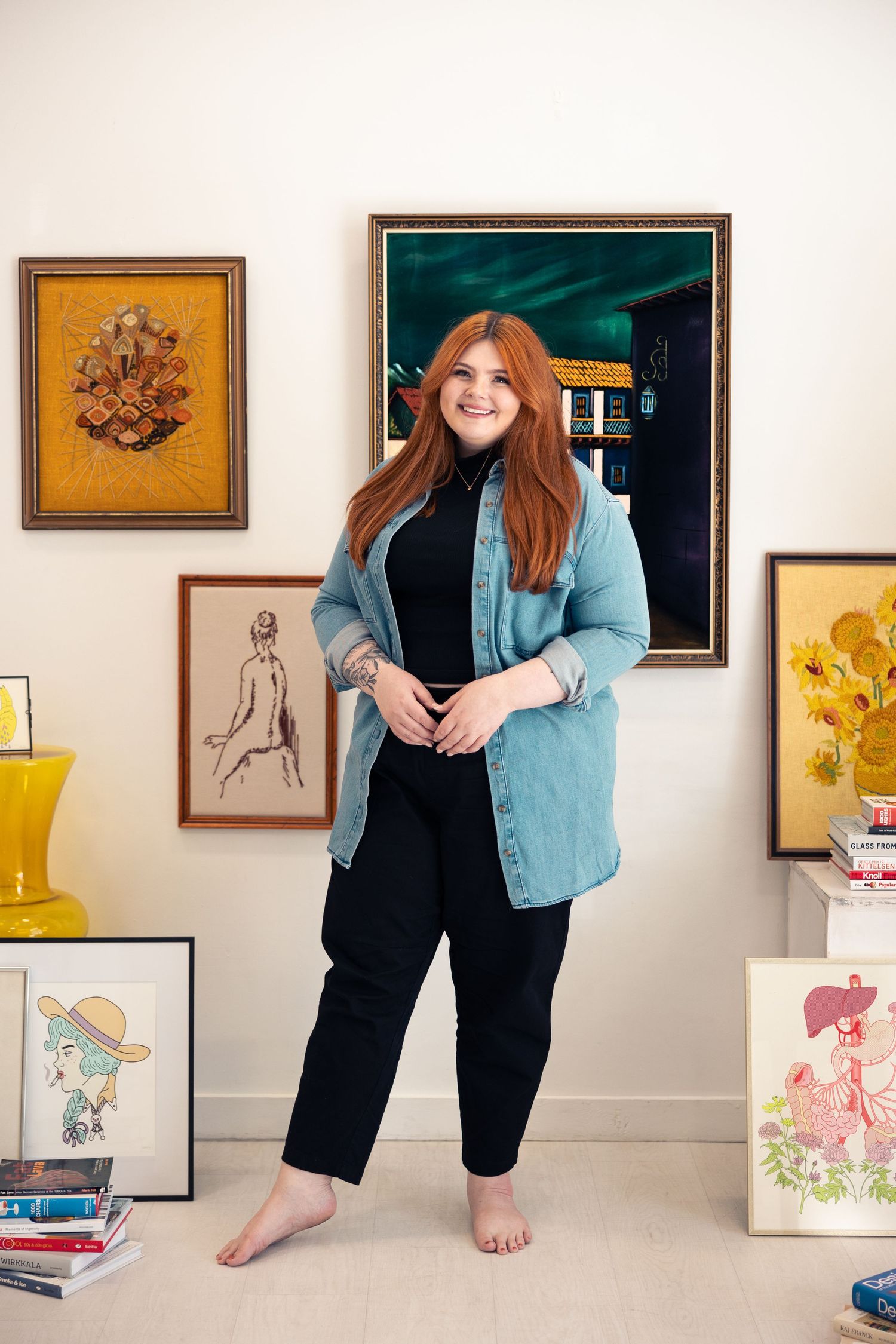 Smiling redhead woman standing barefoot among vintage art collection in bright white room.
