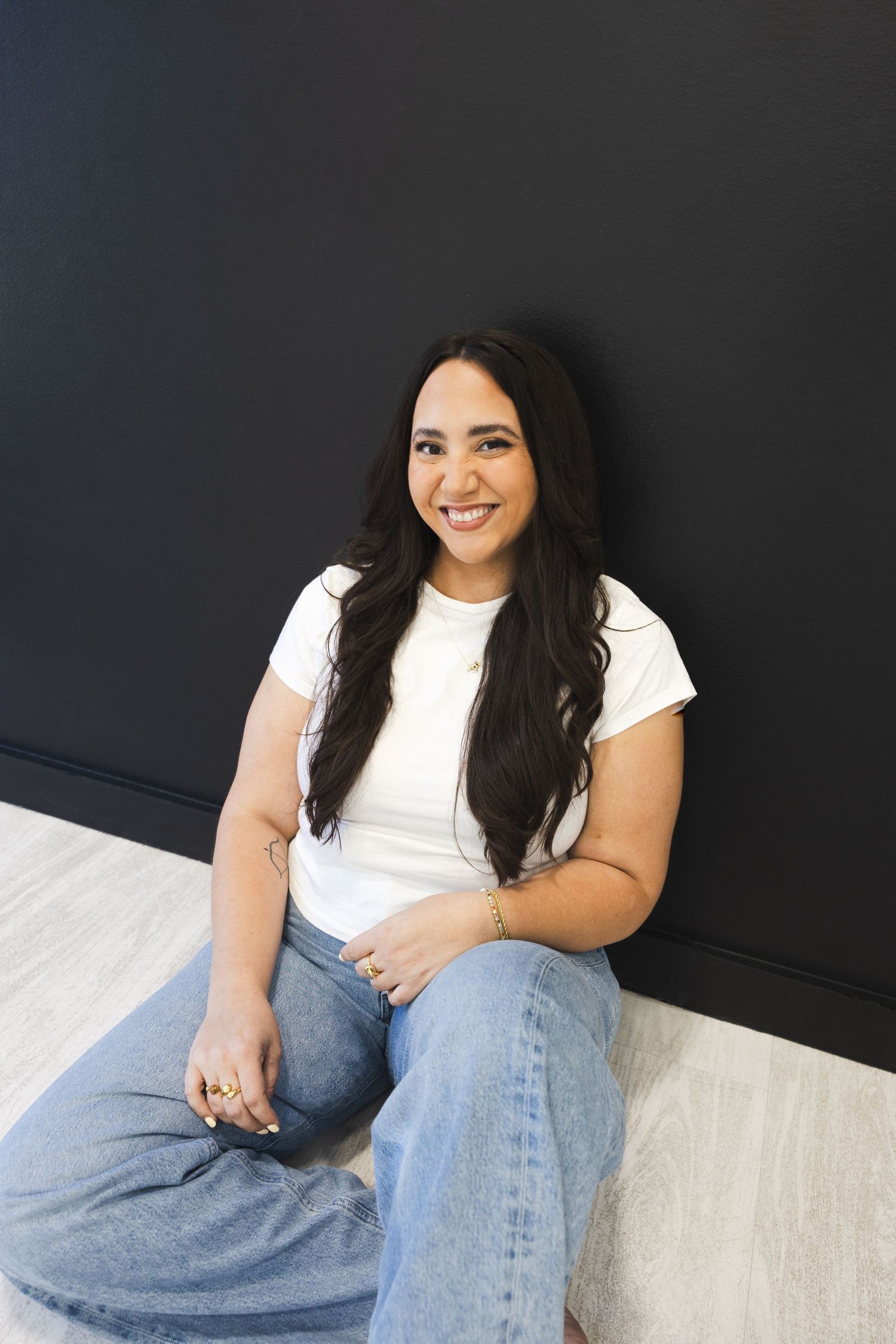 Smiling woman in white tee and jeans sitting on floor against black wall.