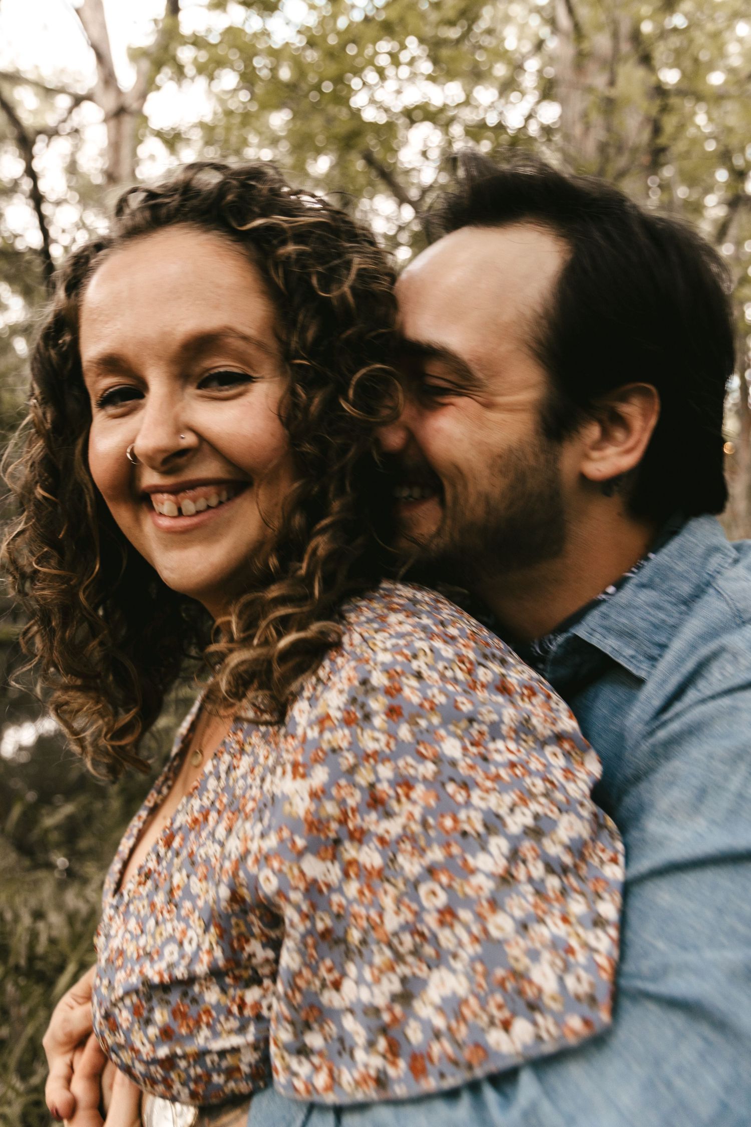 Happy couple embracing in a wooded setting, woman smiling with curly hair in floral dress.