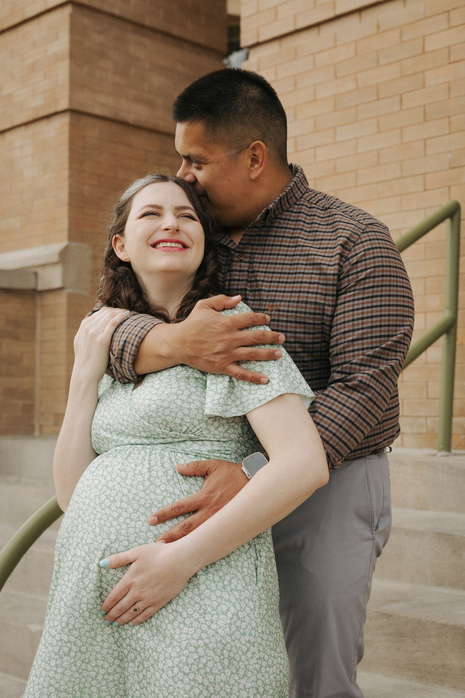 A smiling pregnant woman in a green floral dress being embraced by a man in a plaid shirt on outdoor steps.