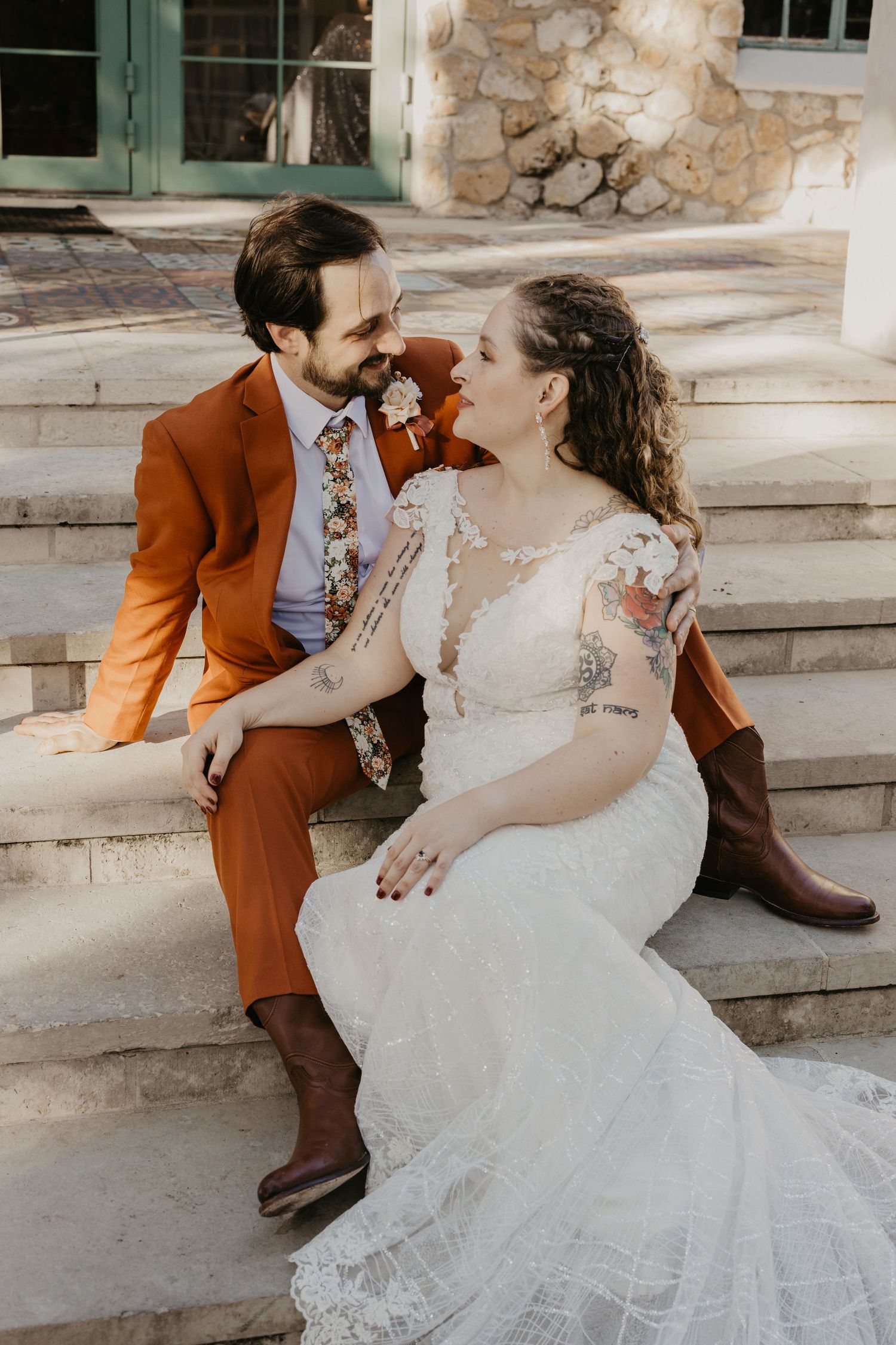 Bride and groom sitting on stone steps, groom in rust orange suit with floral tie, bride in lace wedding dress with cowboy boots.