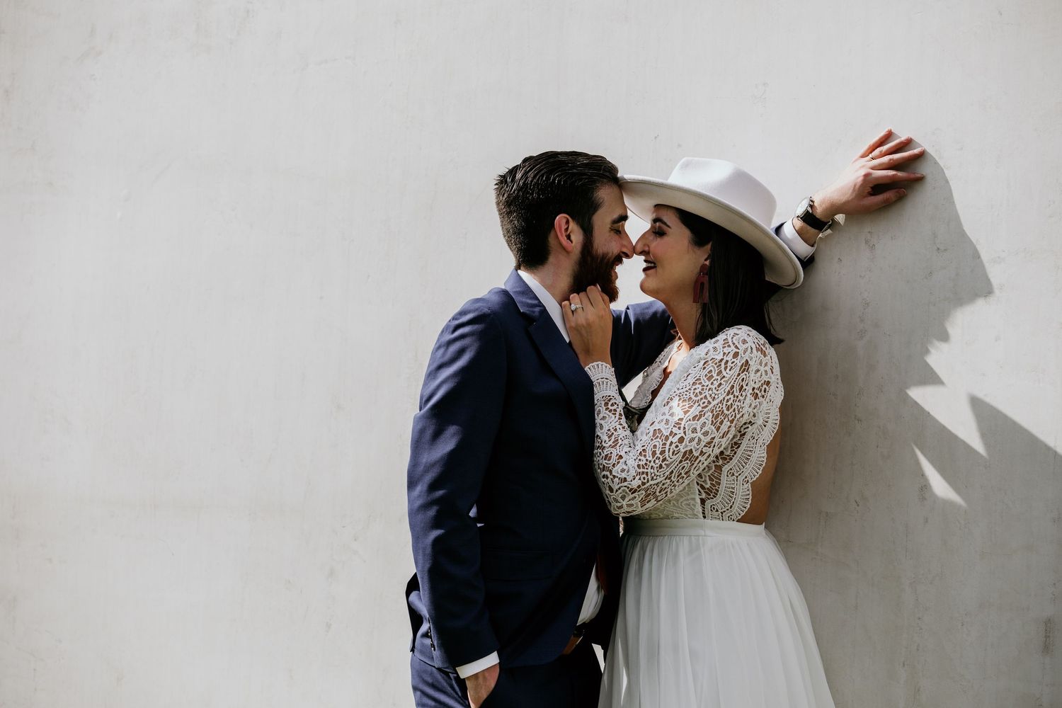A couple in wedding attire shares a romantic moment against a white wall, bride wearing a lace dress and white hat.