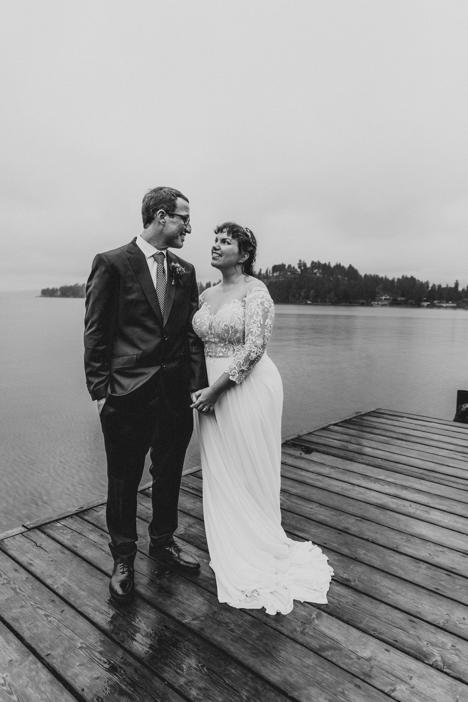 Black and white photo of bride and groom holding hands and smiling on a wooden dock by the water.
