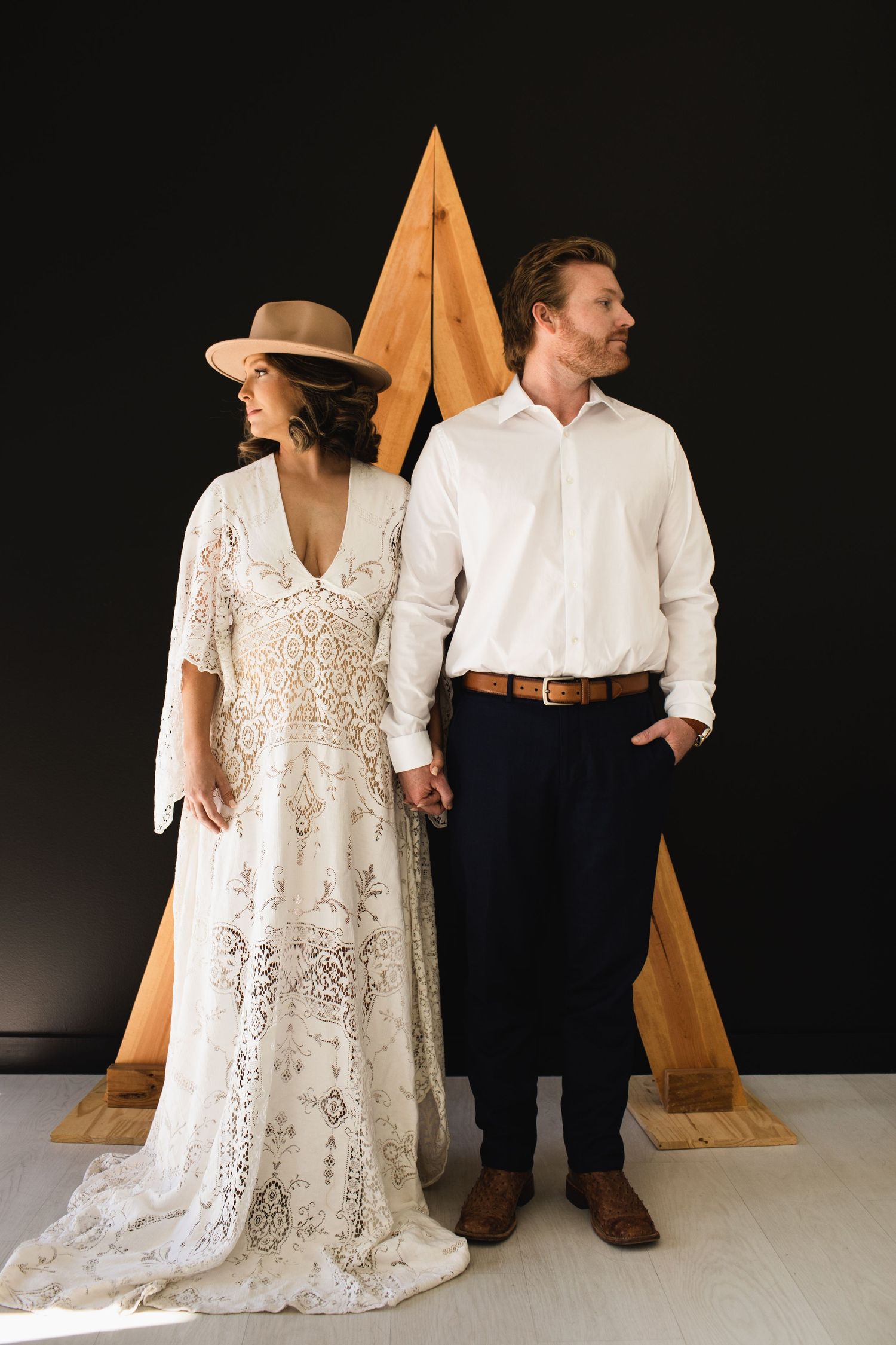Boho bride in lace gown and hat holds hands with groom in white shirt before geometric wood backdrop.