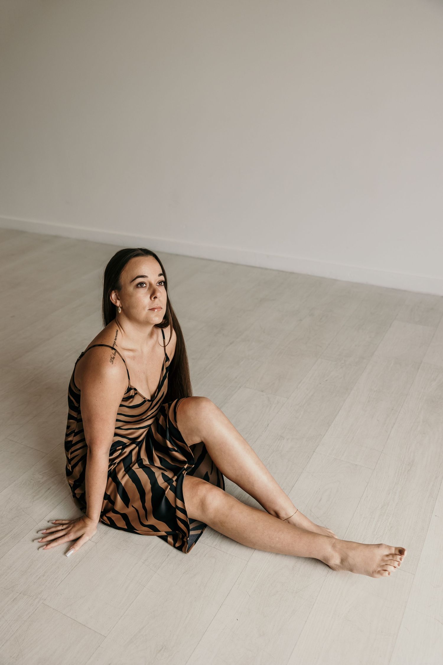 Woman in tiger stripe satin dress sitting barefoot on light wood floor in minimal studio.