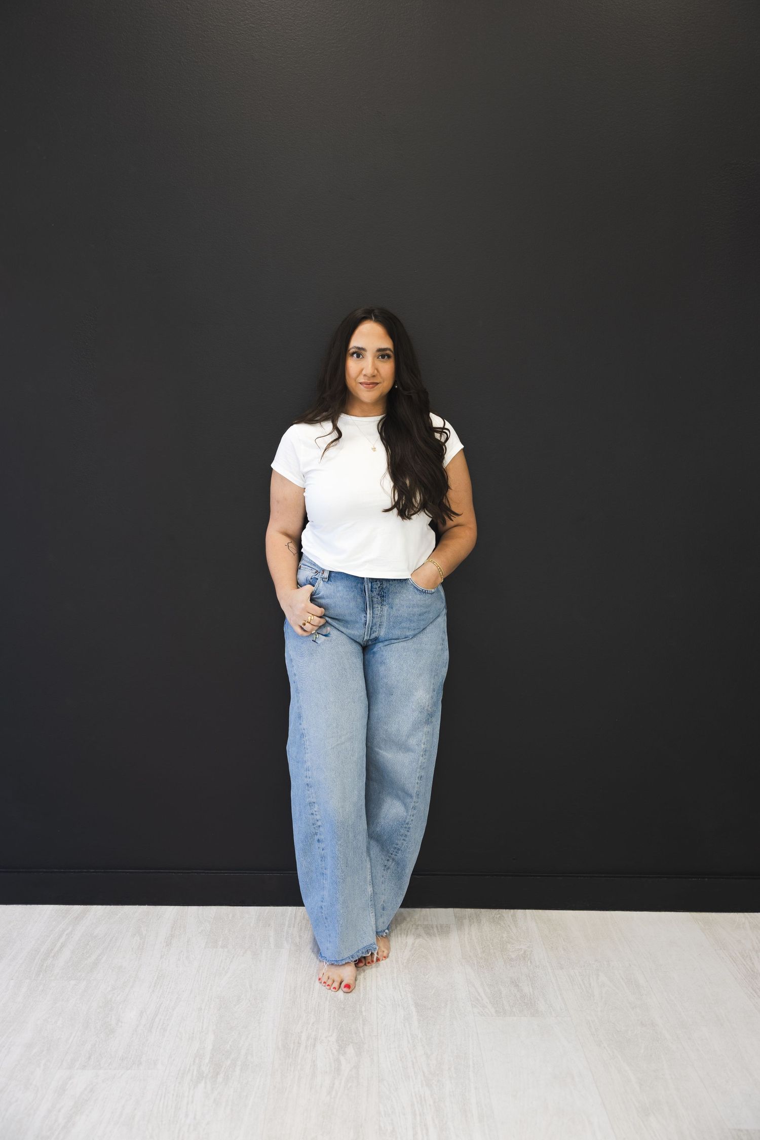 Woman in white tee and wide-leg jeans standing against dark gray wall.