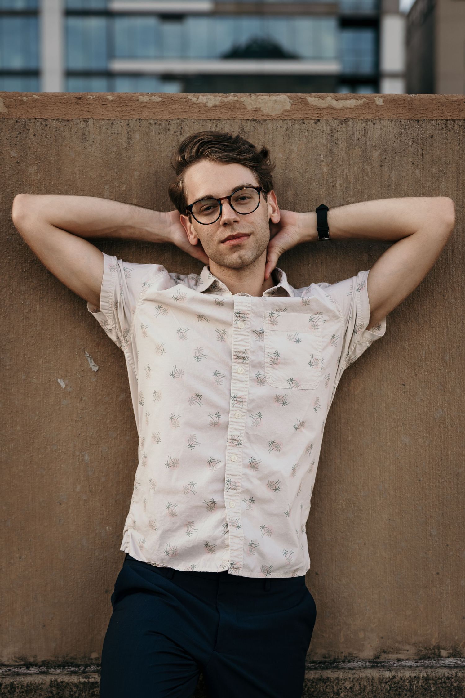 Young man in a floral short-sleeve shirt relaxes with hands behind head against a concrete wall.