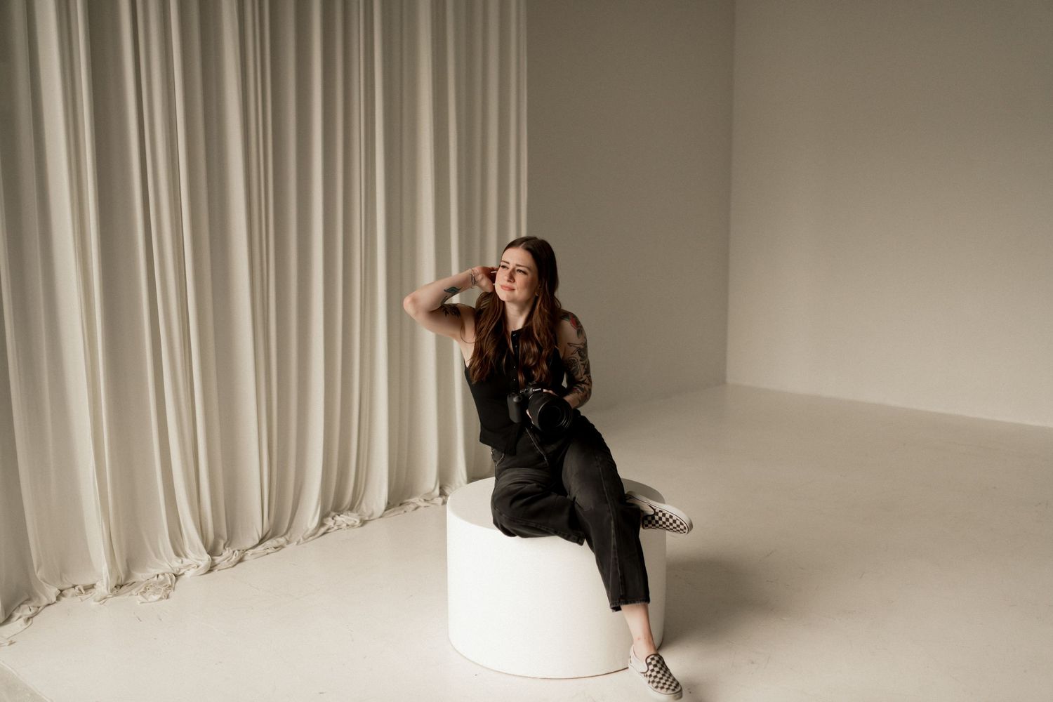 Tattooed female photographer sitting gracefully on white pedestal in black outfit against neutral studio backdrop.